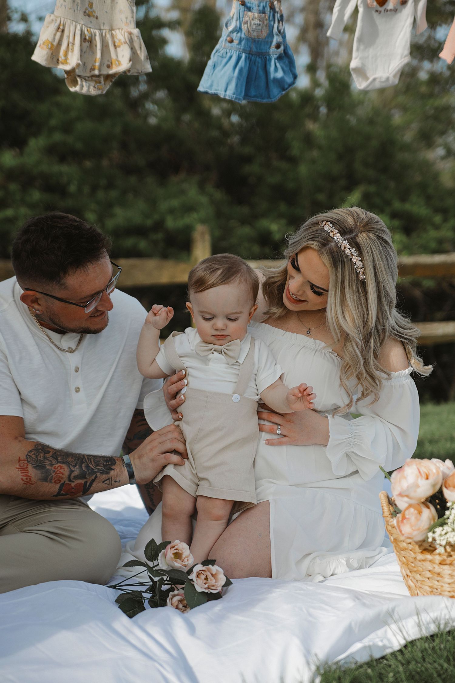A series of tender family moments captured on a white blanket in a grassy outdoor setting with hanging vintage clothing above.