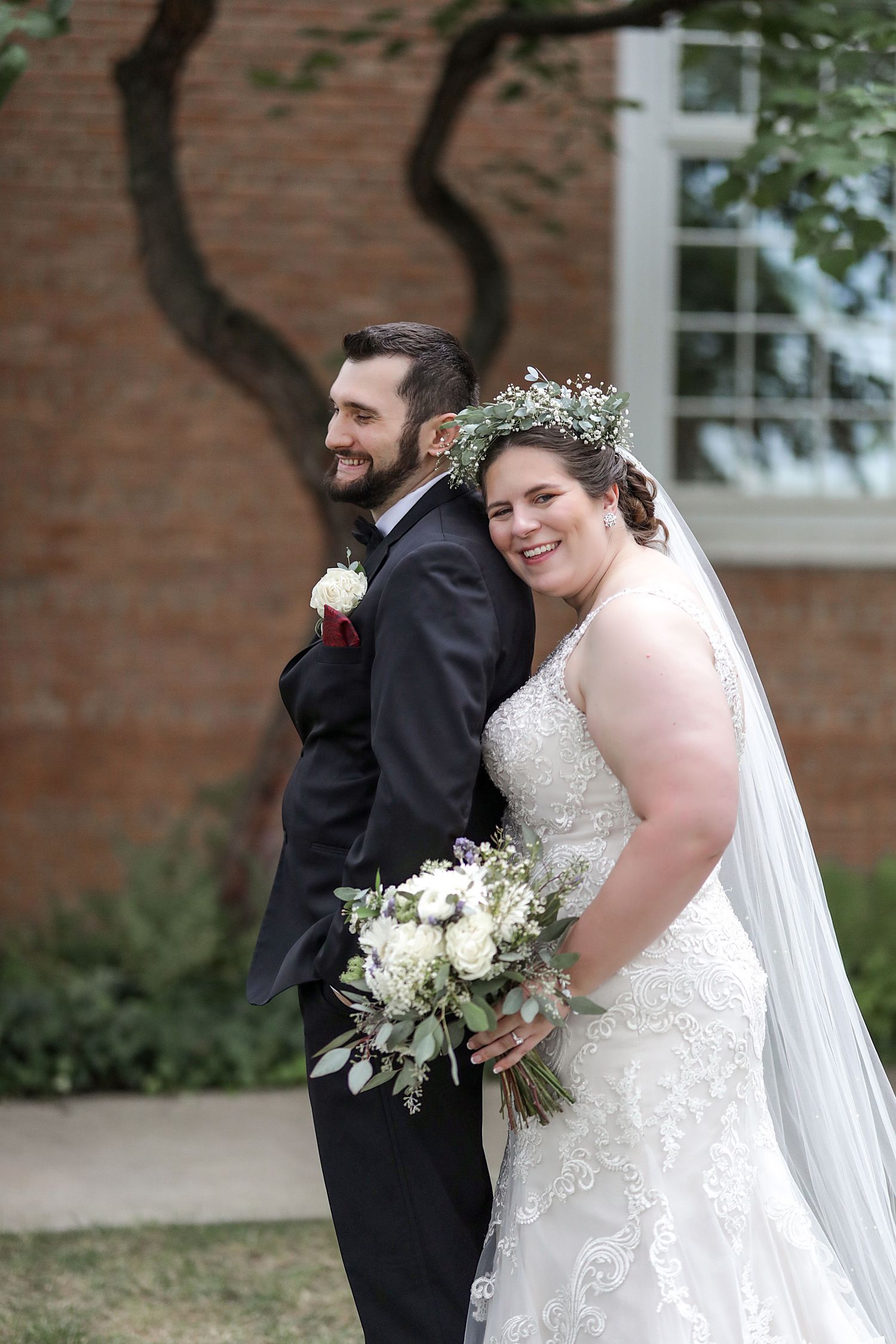 A happy couple poses together at their wedding, wearing traditional bridal attire and a floral crown against a brick backdrop.