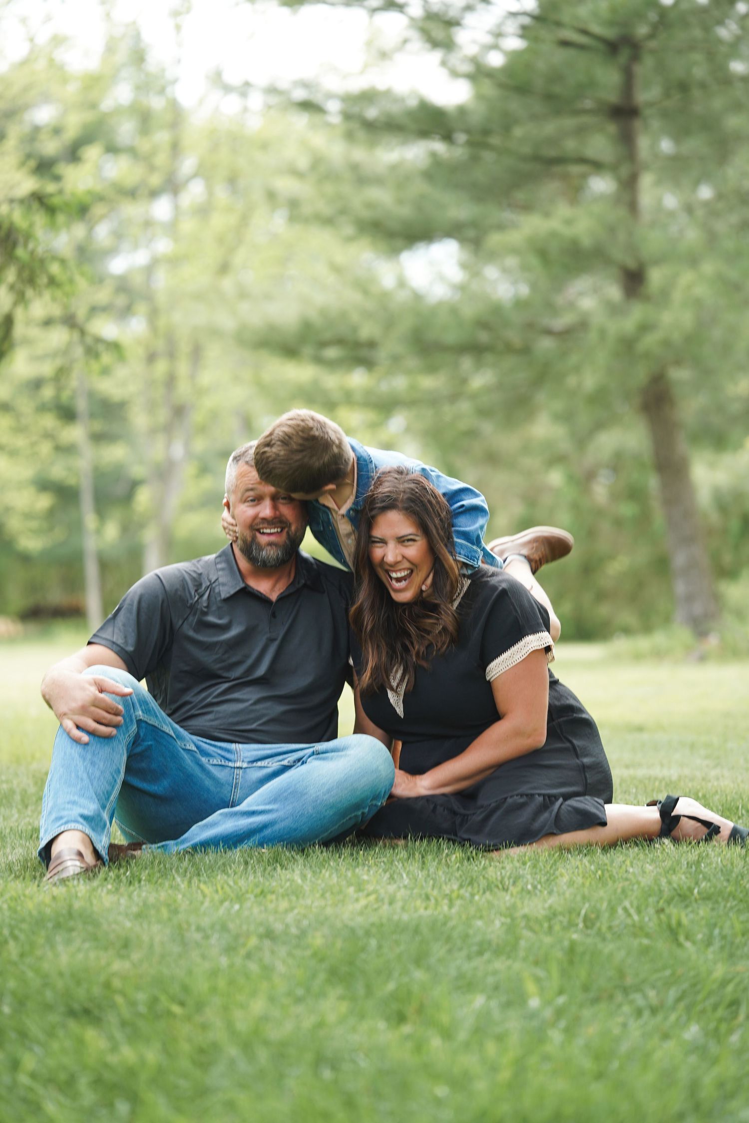 A couple sits together and laughs on green grass in a wooded outdoor setting during a casual photo session.