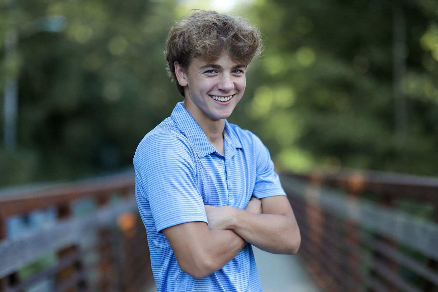 Young teen in light blue polo shirt stands with arms crossed and a bright smile outdoors on a wooden bridge.