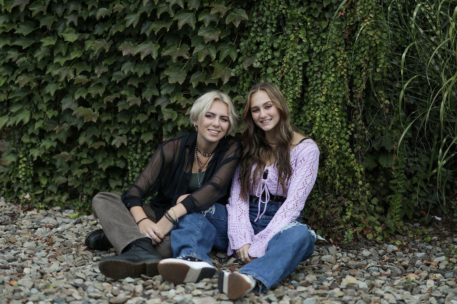 Two people sit together on stone ground against a lush green ivy-covered wall outdoors.