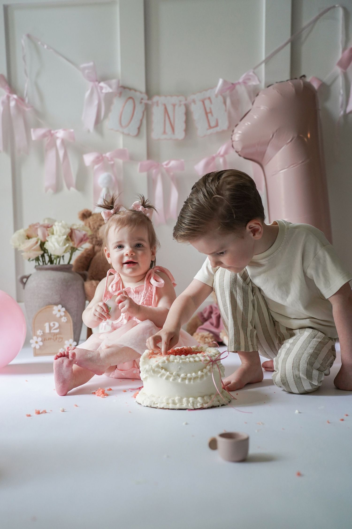 Baby smashing cake during first birthday cake smash photo session with pink decorations and garland in background.