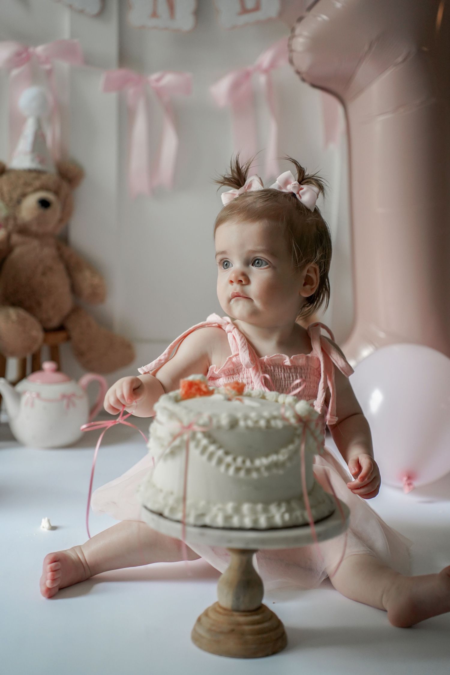 A series of photos show a baby's first birthday cake smash session with a white frosted cake on a vintage pedestal stand.