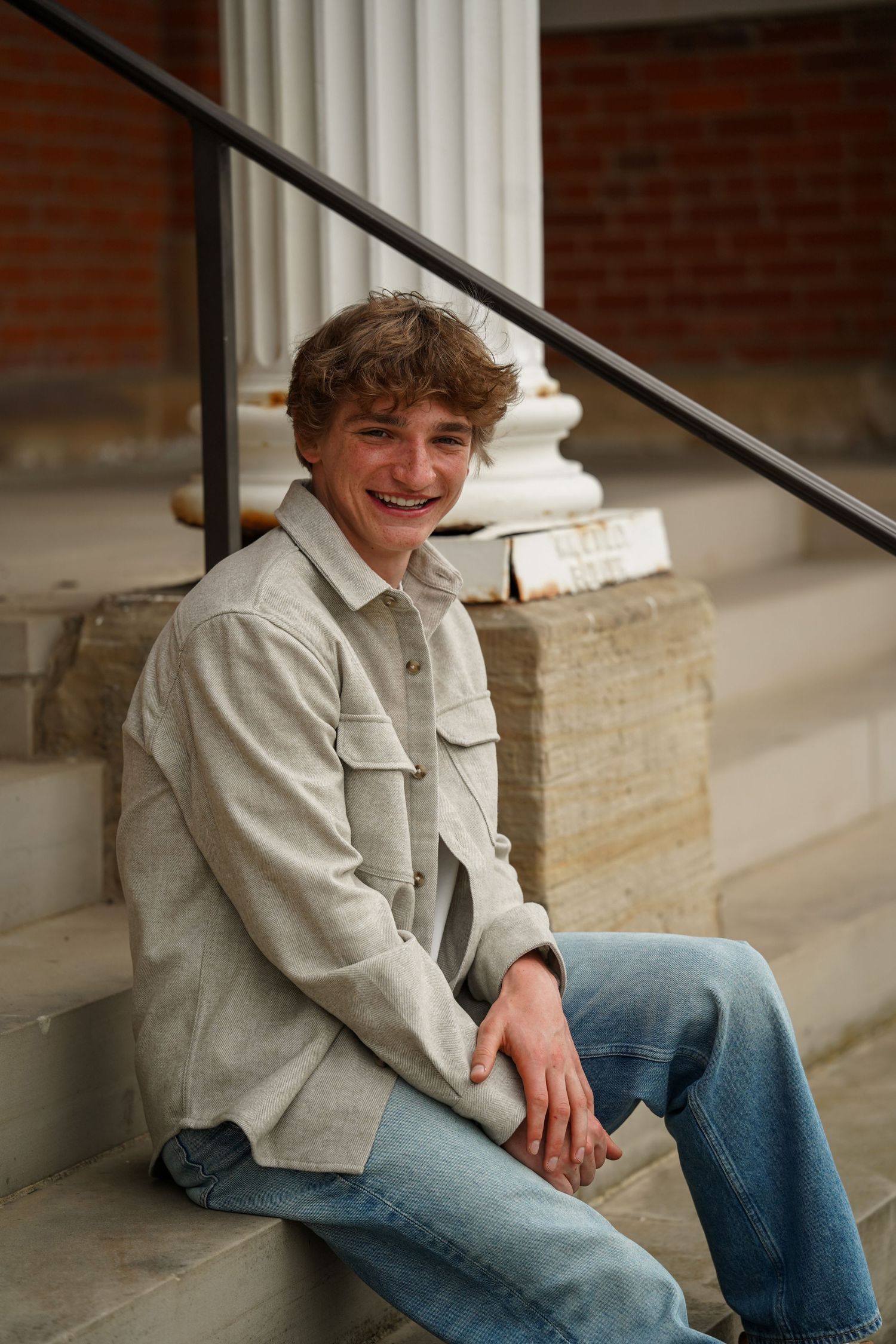 Young person sitting on stone steps wearing a beige jacket and jeans in front of a brick building.