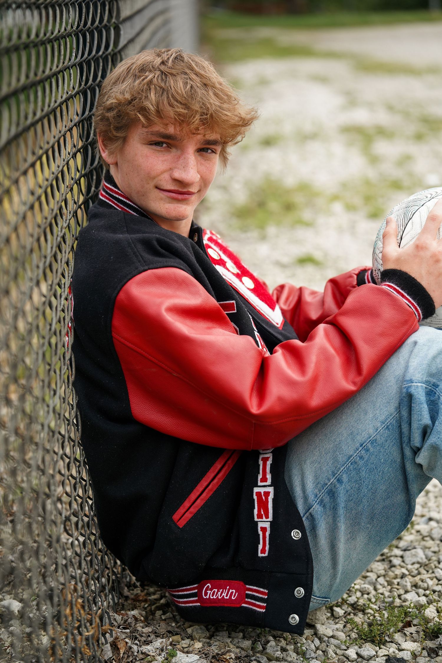A person wearing a red and black varsity jacket leans against a chain link fence while holding a ball.