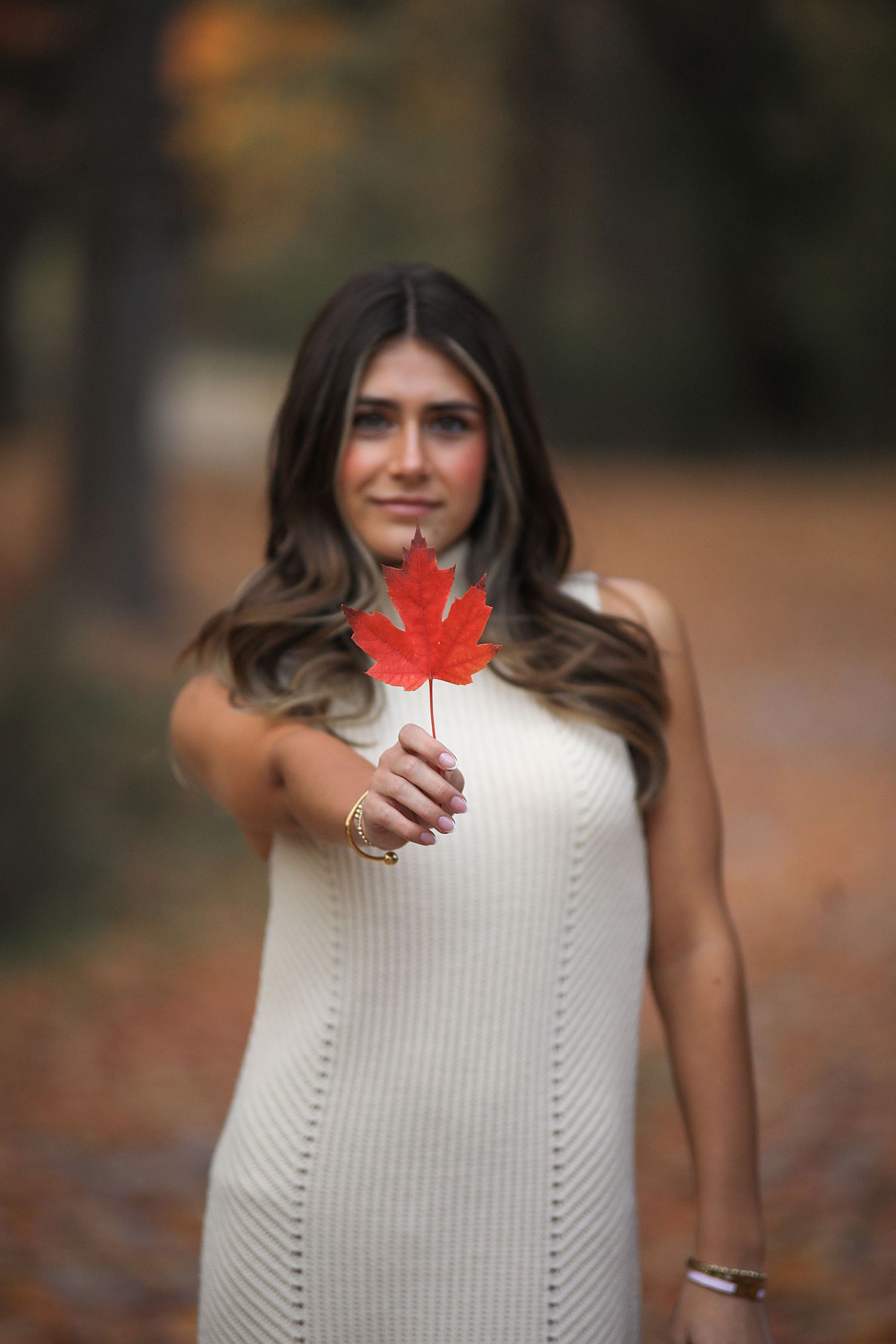 Someone in a white sweater holds a bright red maple leaf in autumn outdoors during a photoshoot.
