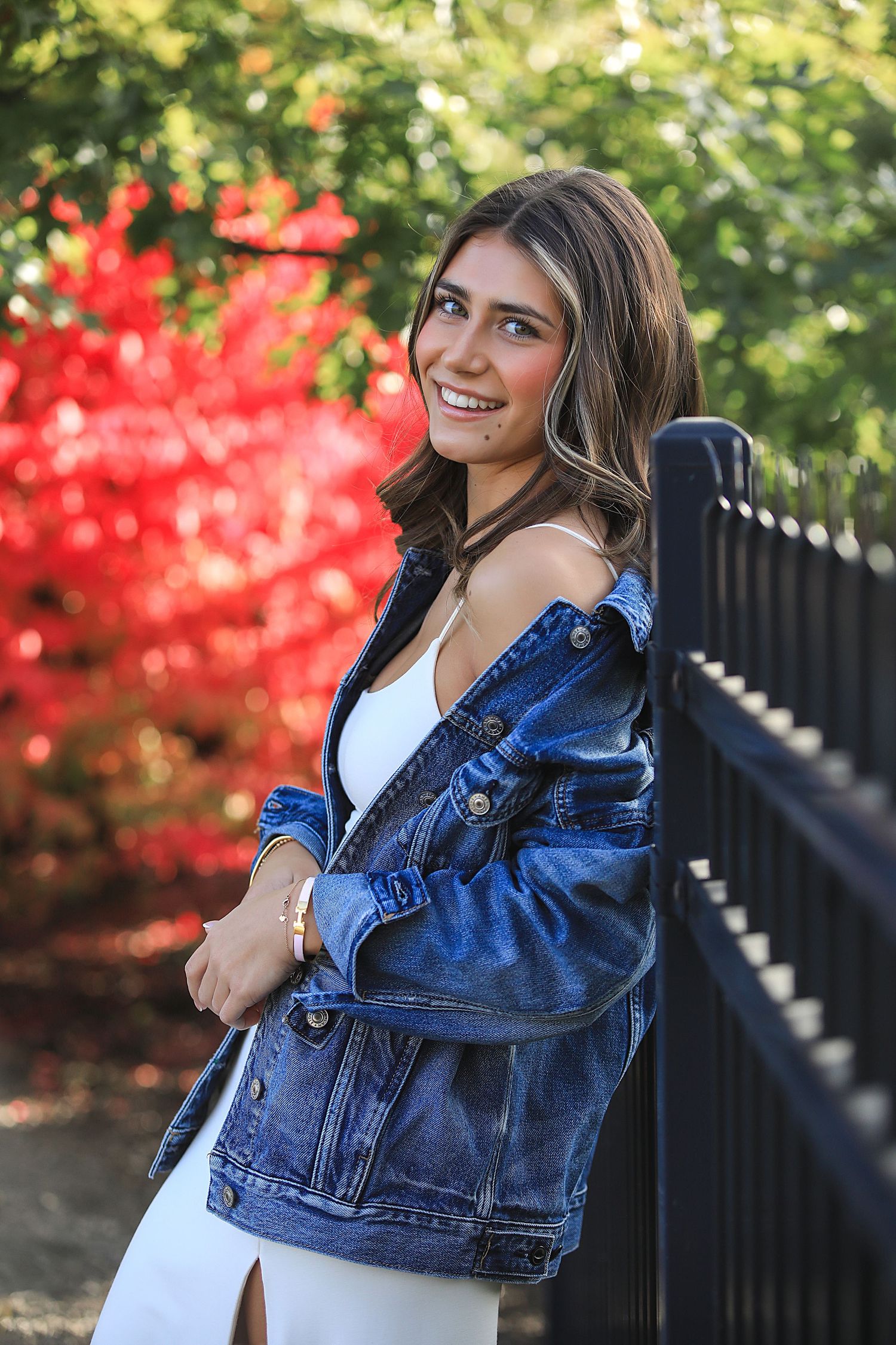 Stylish denim jacket paired with white dress against autumn foliage with bright red leaves in background on sunny day.