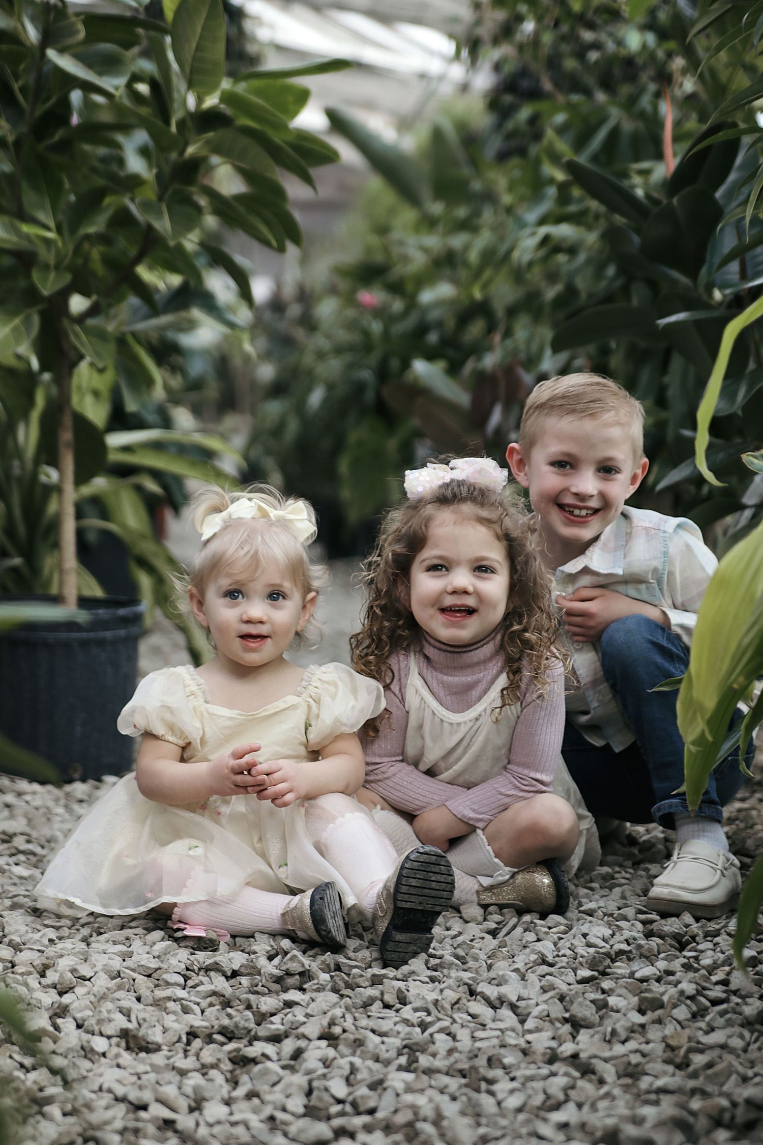 Children sit and play together on decorative gravel surrounded by lush green plants in a greenhouse setting.