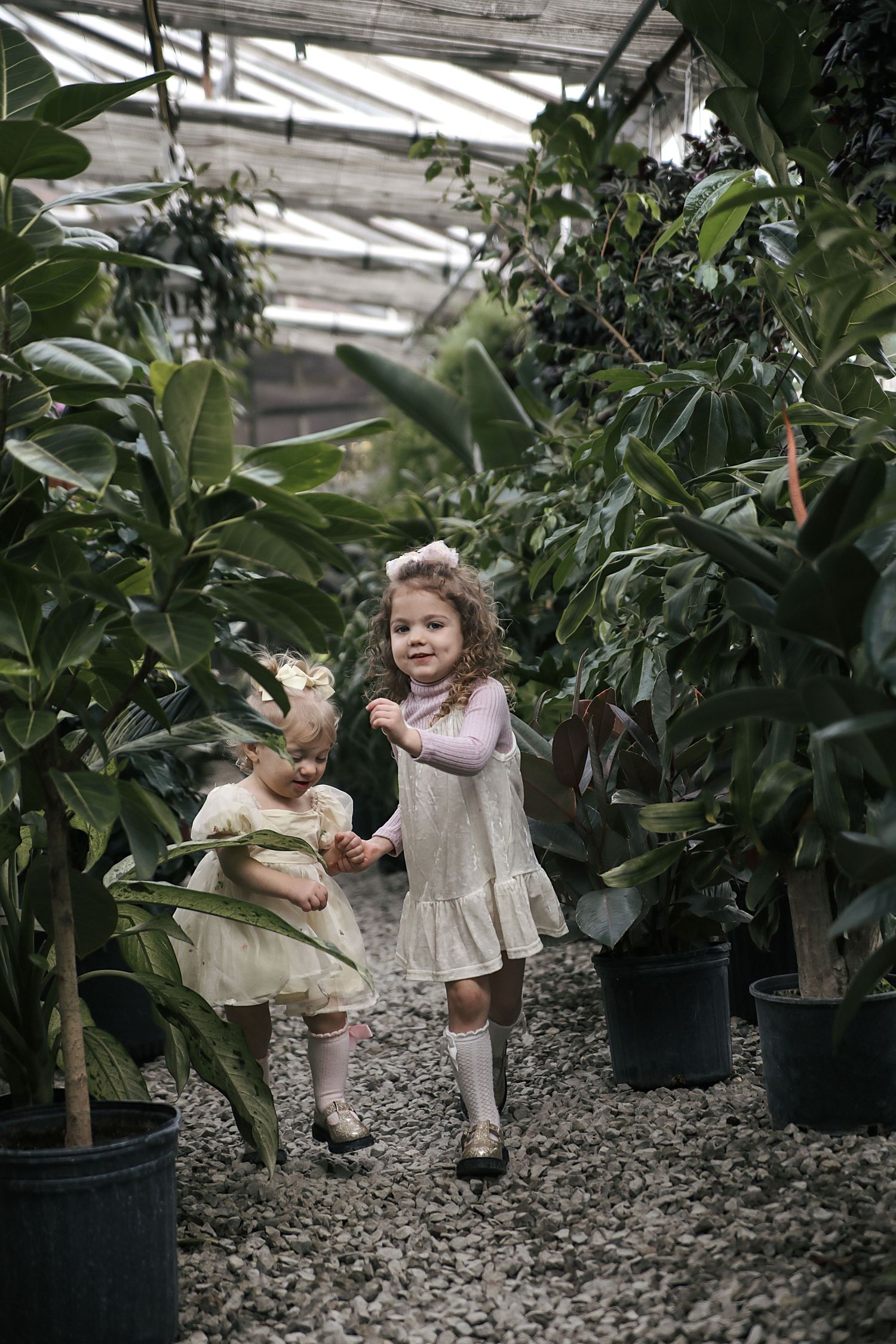 Small children in vintage-style dresses play among tall potted plants in a greenhouse setting.