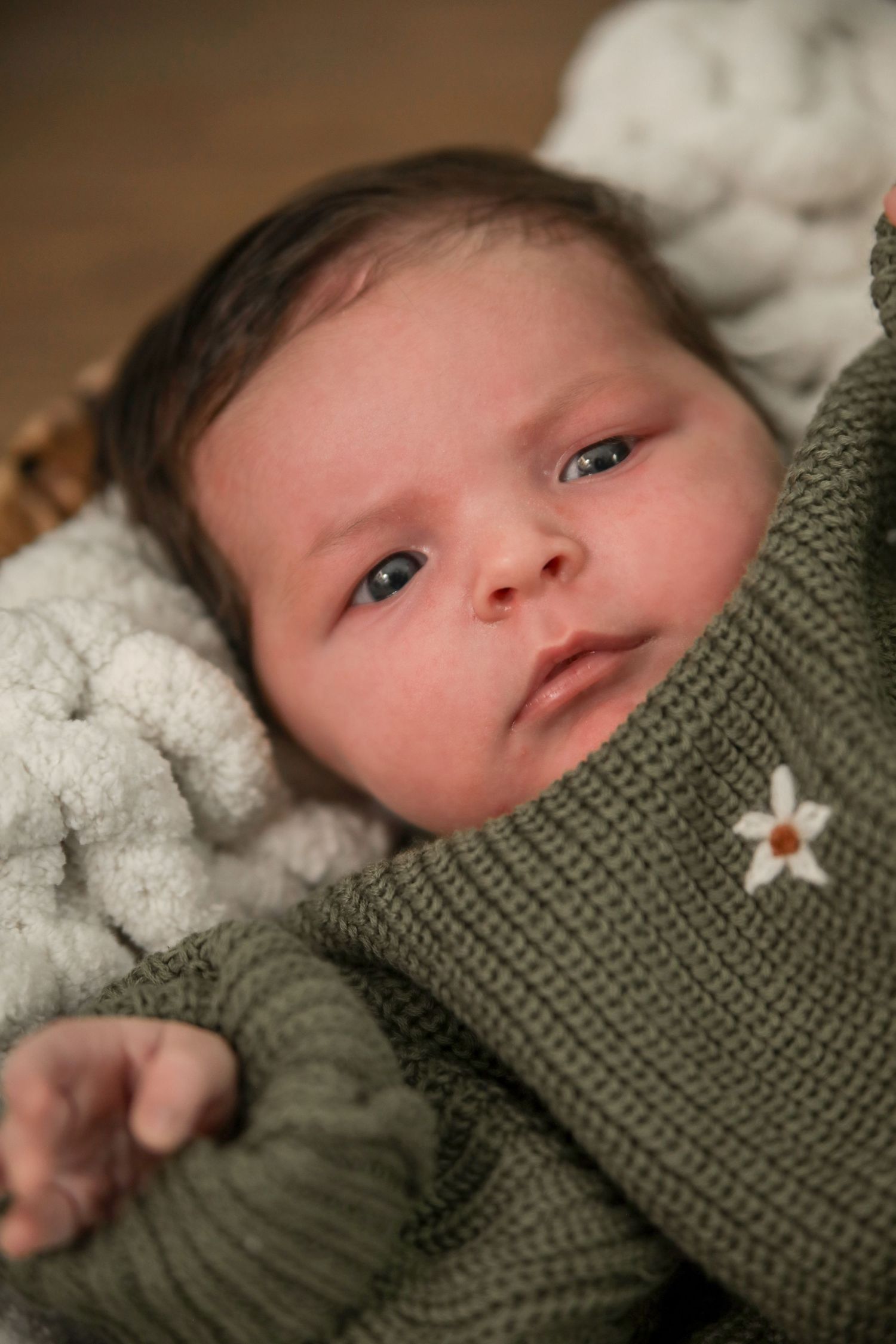 A baby in a green knit sweater lies on soft white bedding while gazing upward with bright eyes.