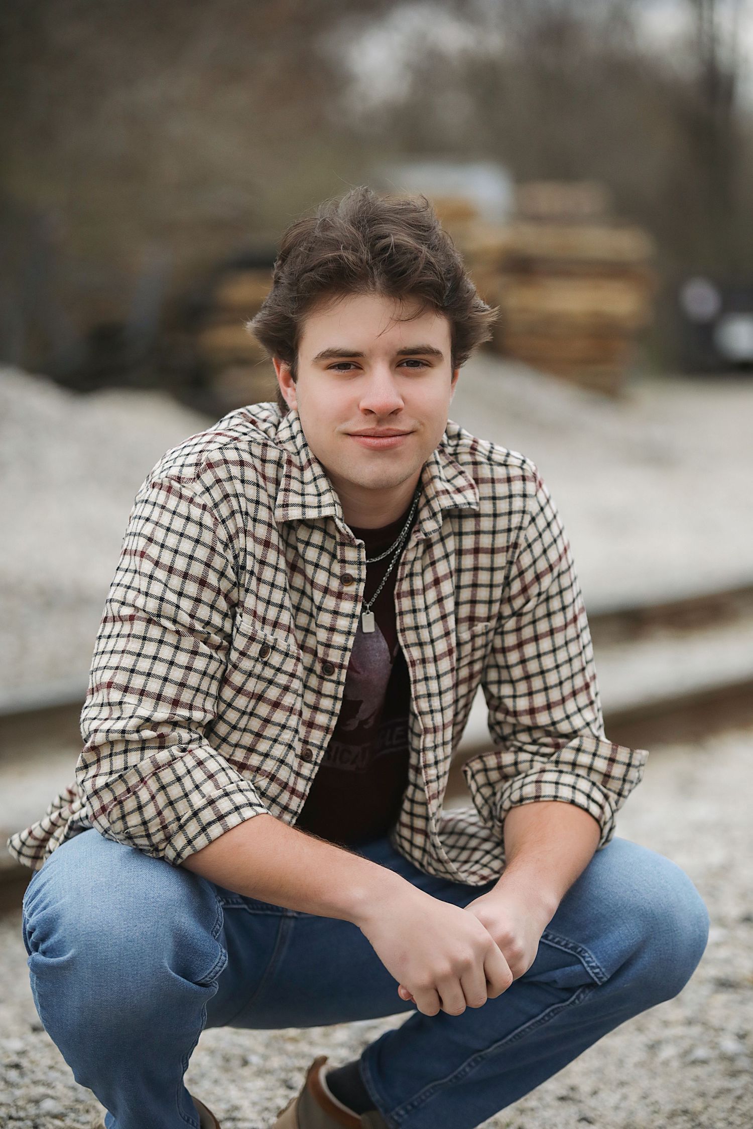 A young person in a plaid button-up shirt and jeans crouches outdoors near stacked lumber on an overcast day.