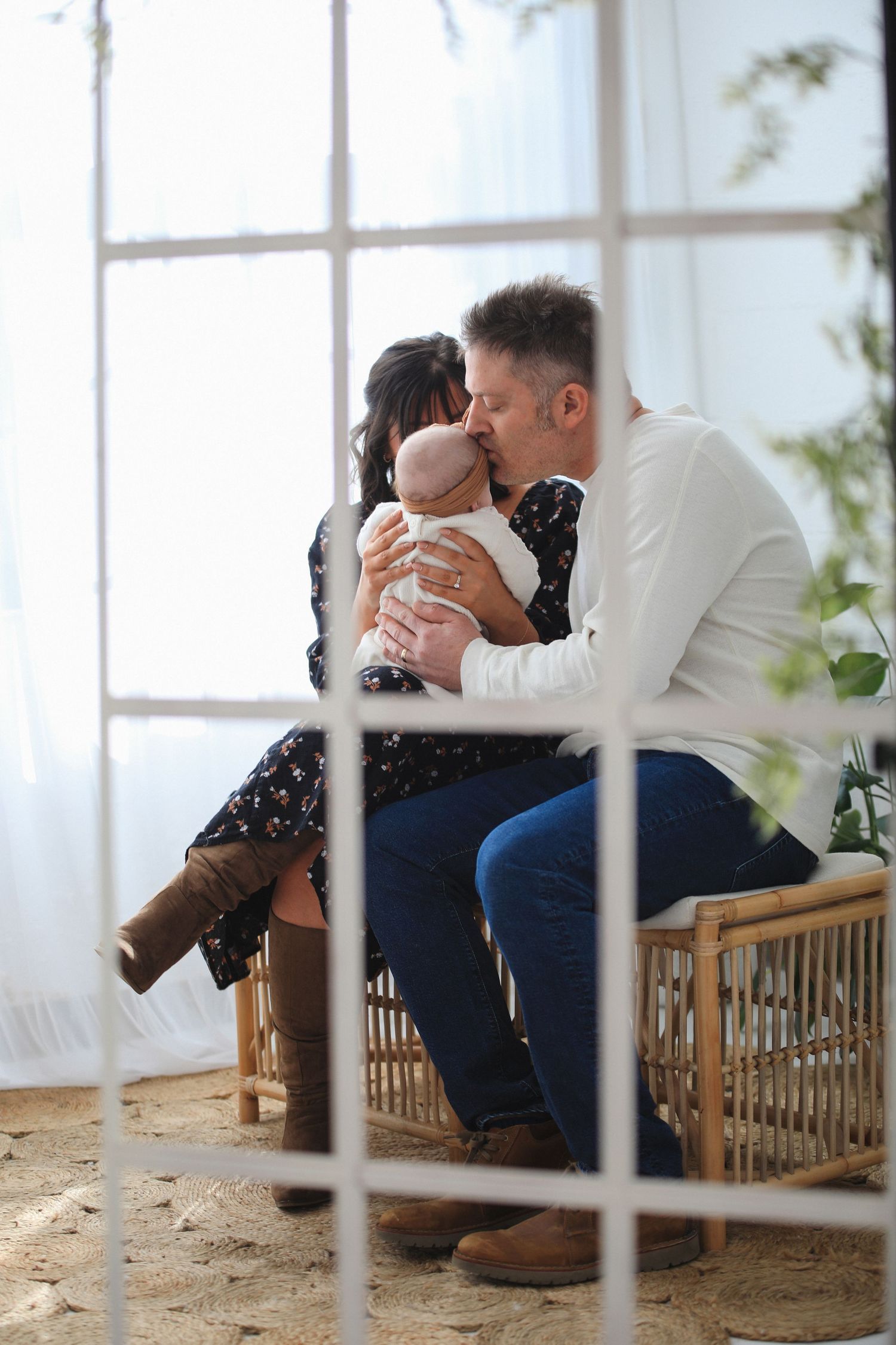 A couple shares an intimate moment while sitting on a wooden bench seen through window panes in a bright room.