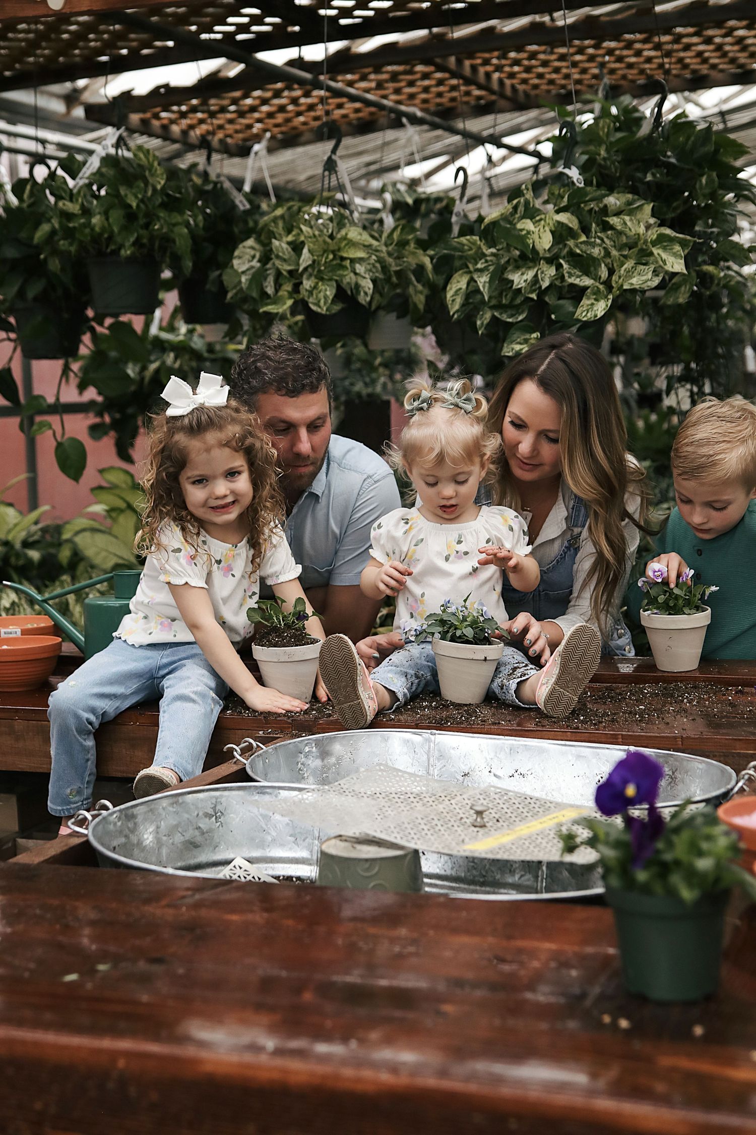 A family plants together at a rustic wooden potting table surrounded by lush green plants and gardening supplies.