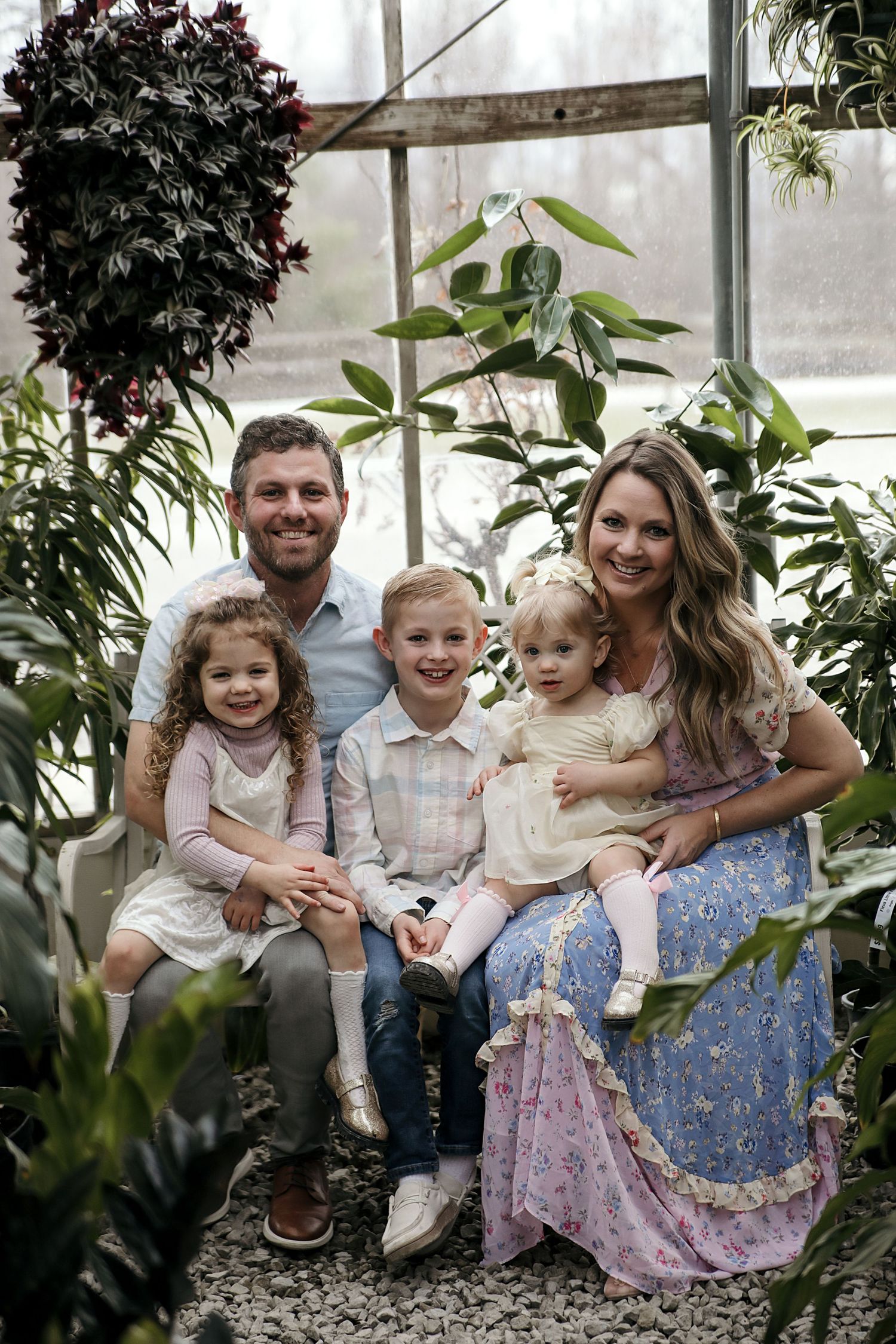 A family sits together on stone steps in a greenhouse surrounded by lush plants wearing casual spring attire.