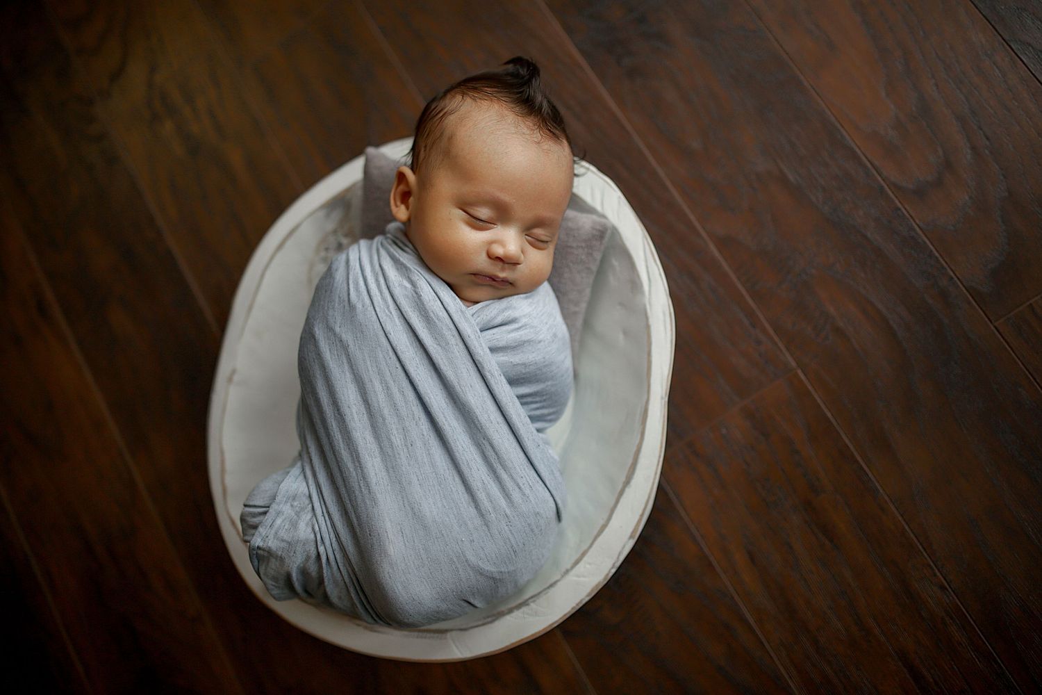 Peaceful sleeping newborn wrapped in soft gray swaddle in white bowl on wooden floor. Newborn photographer Medina, Ohio.