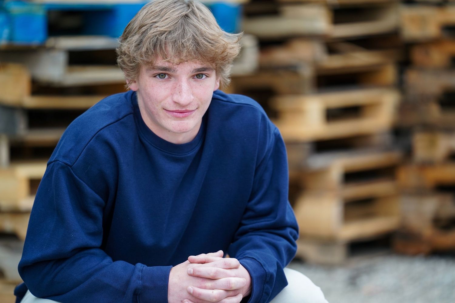 A person in a navy blue sweater is sitting at a wooden table next to pallets stacked in the background.