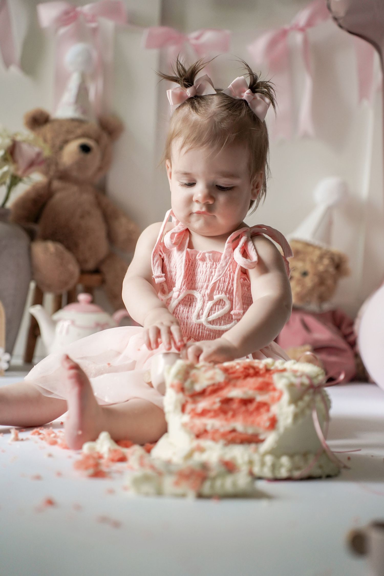 A baby in pink dress delightfully plays with a smashed cake during their first birthday cake smash photoshoot.