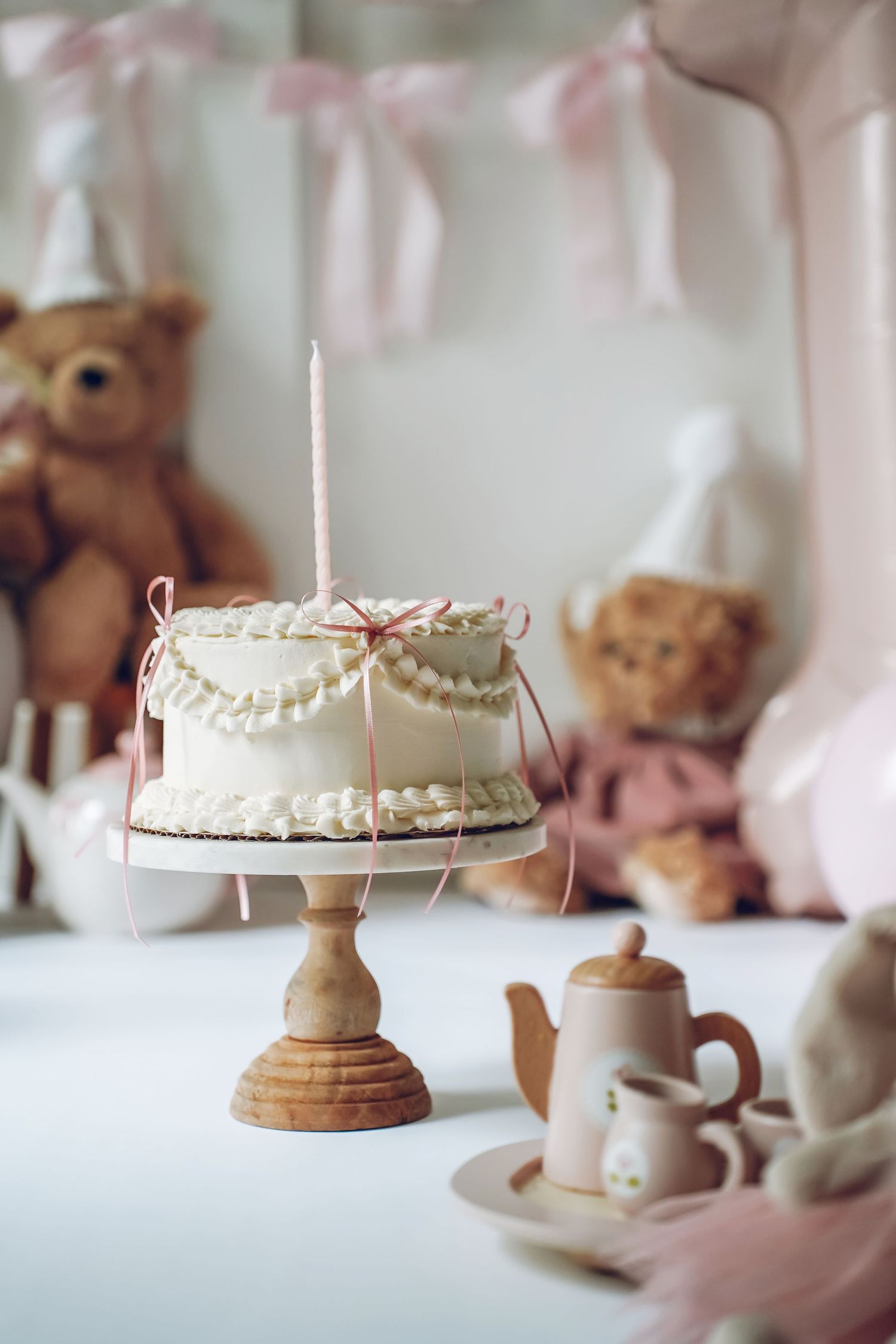 White frosted cake on wooden pedestal with pink ribbons and teddy bears in soft focus background with tea set.