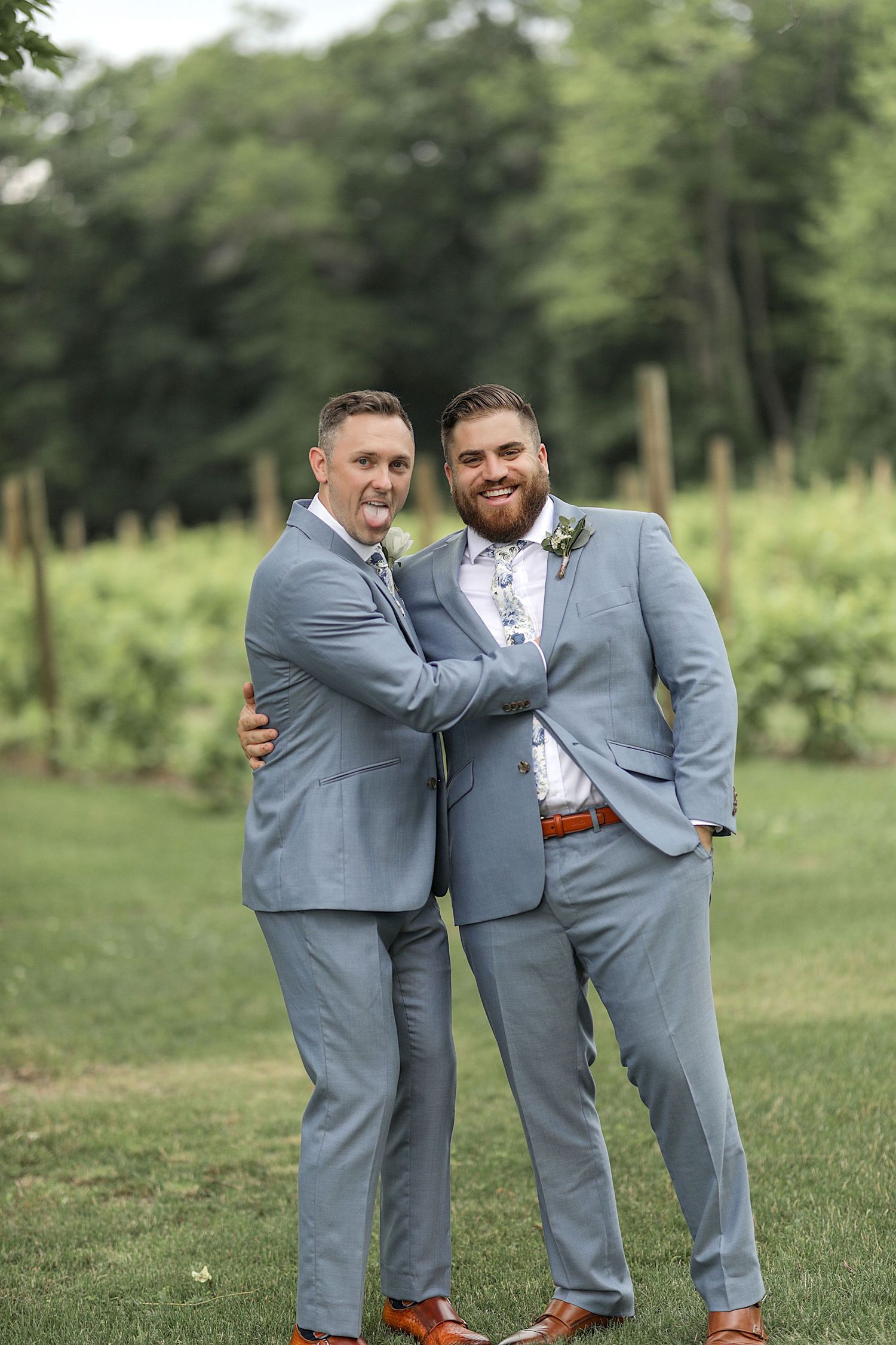Two groomsmen in matching grey suits share a cheerful moment on the lawn.