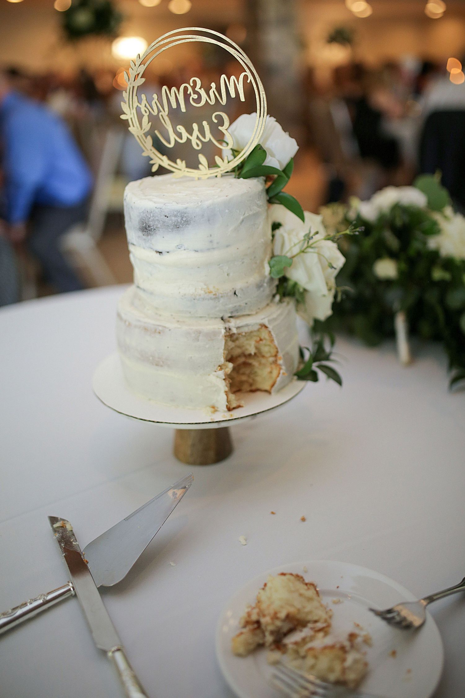A semi-naked wedding cake with white florals and a gold topper on display.
