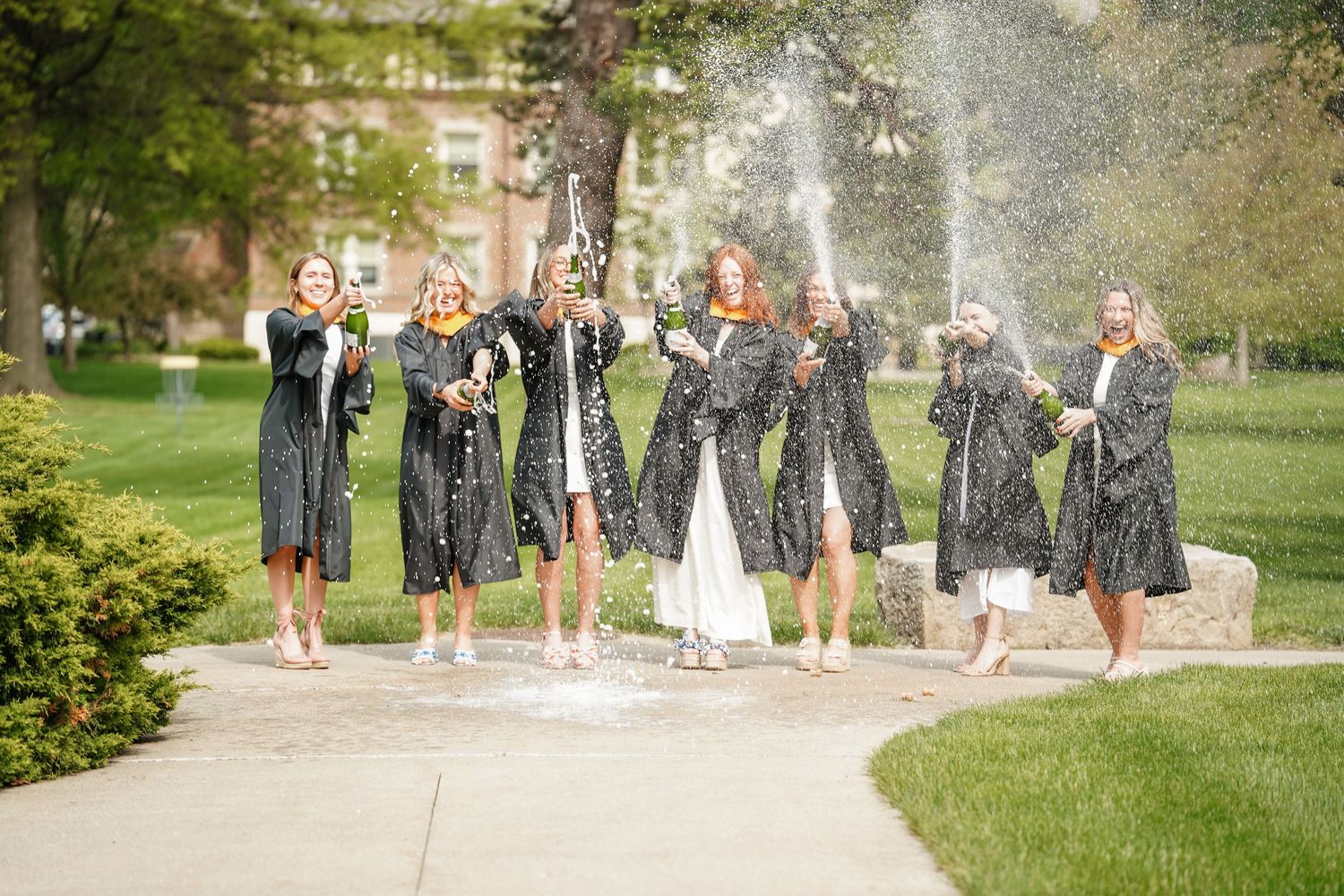 A joyful wedding party in navy dresses spraying champagne on a sunny garden path among green landscaping.