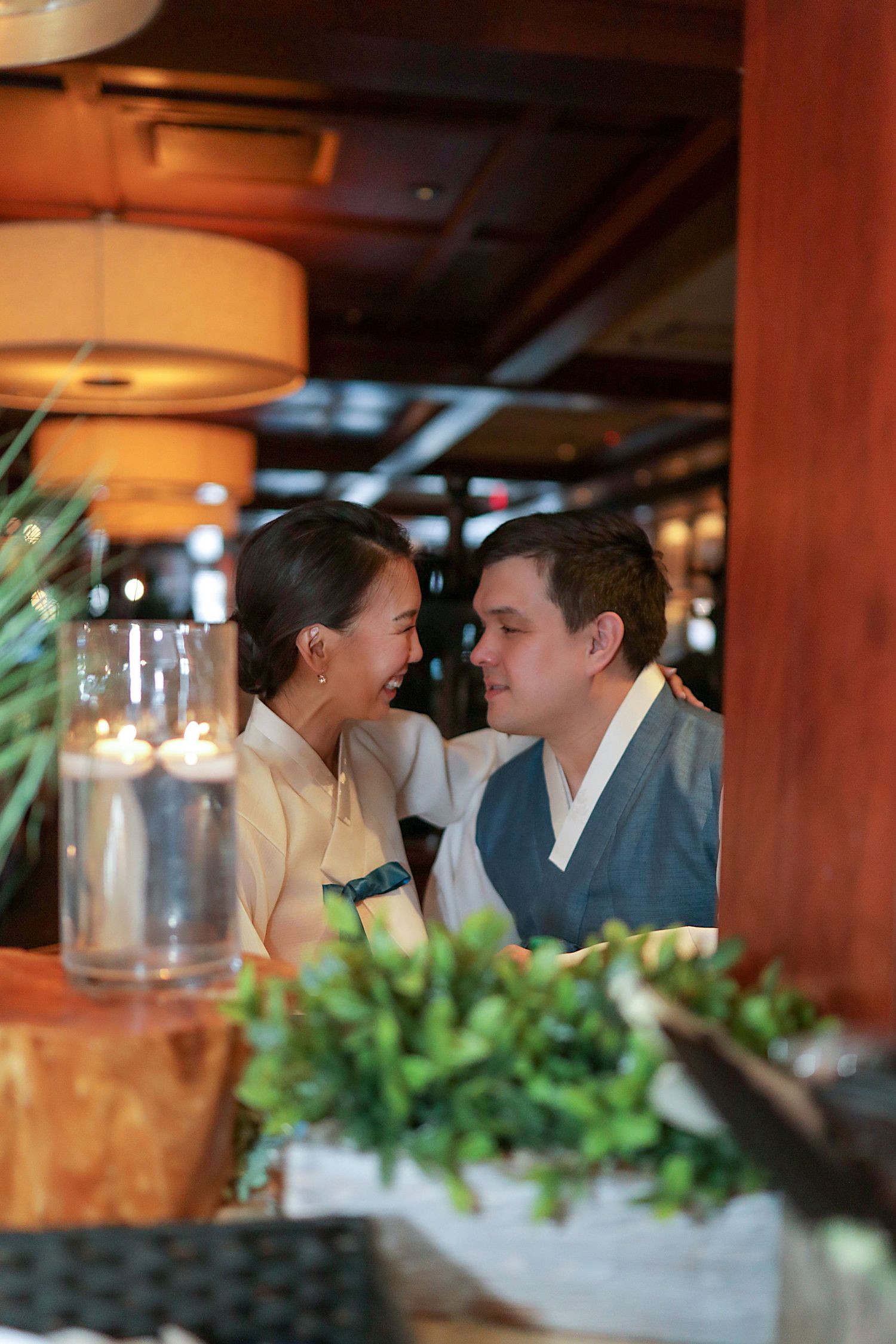 A couple shares an intimate moment at a dimly lit restaurant table with wine glasses and greenery in the foreground.