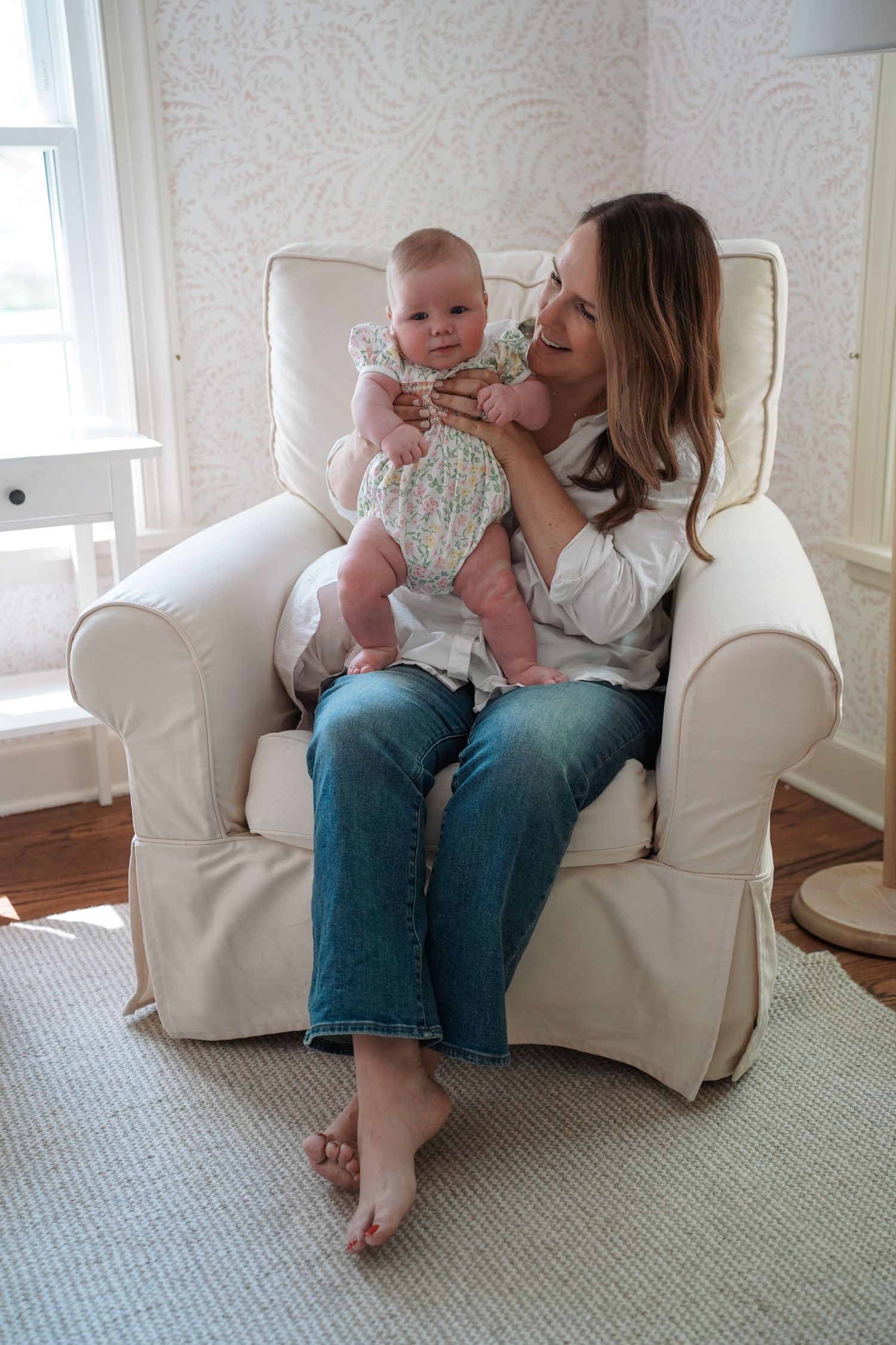 Two people share a tender moment while sitting in a cozy white armchair against a paisley wallpapered wall.