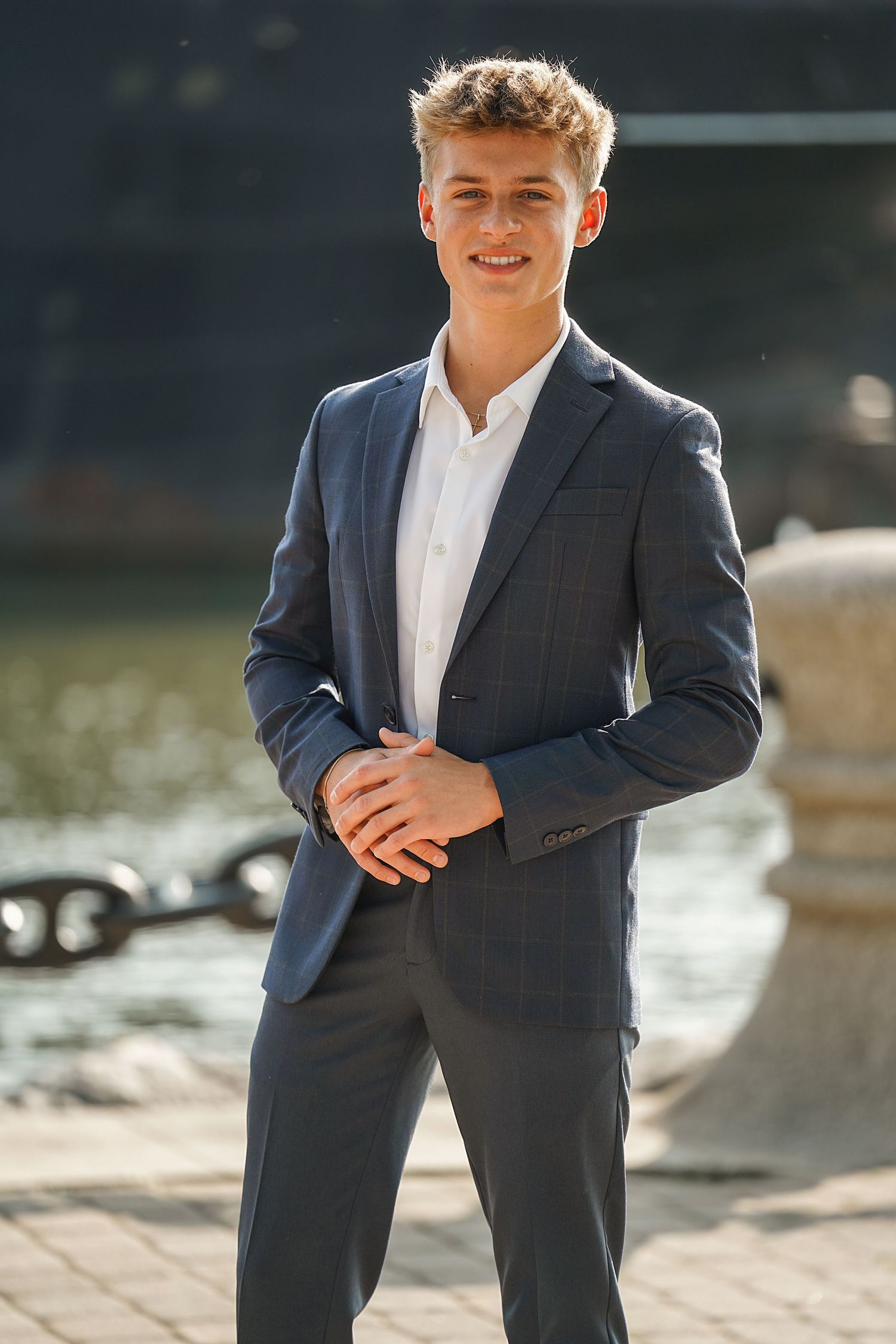 A person standing outdoors in a navy business suit and white button-down shirt poses against a waterfront background.