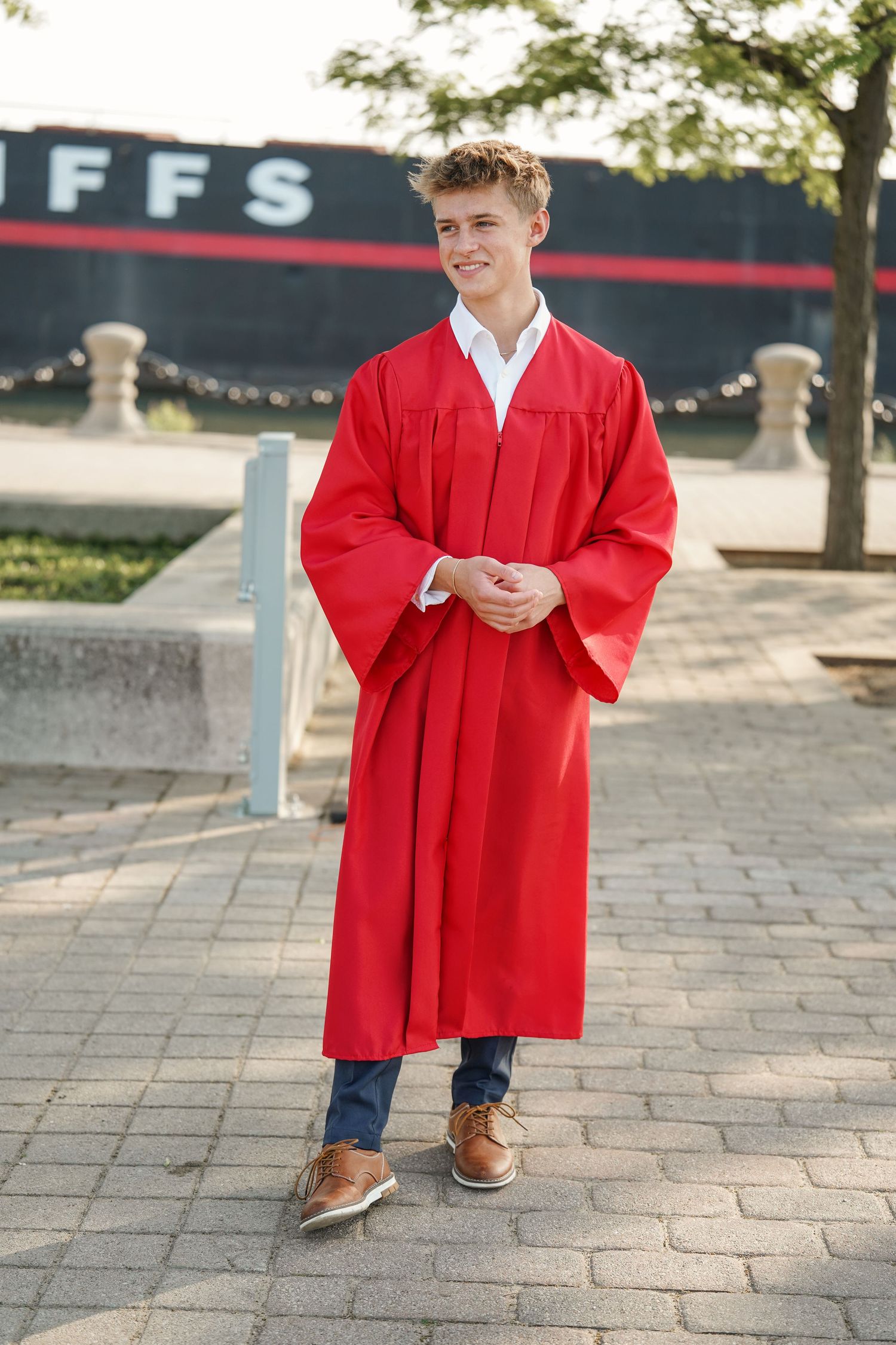 Graduate in red cap and gown poses outdoors on brick walkway wearing brown leather shoes.