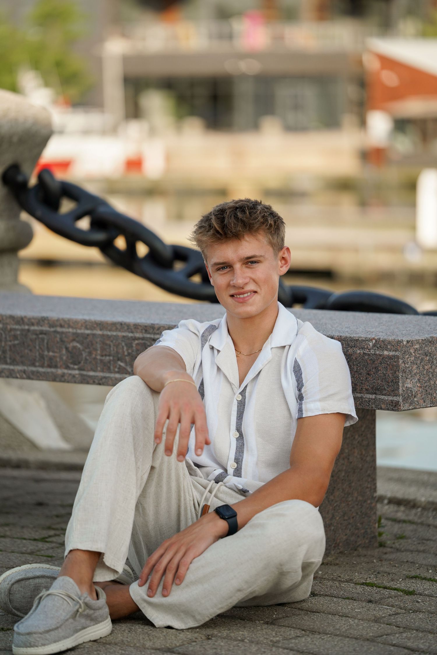 Young person wearing casual white top and light pants poses while sitting on stone ledge near waterfront area.