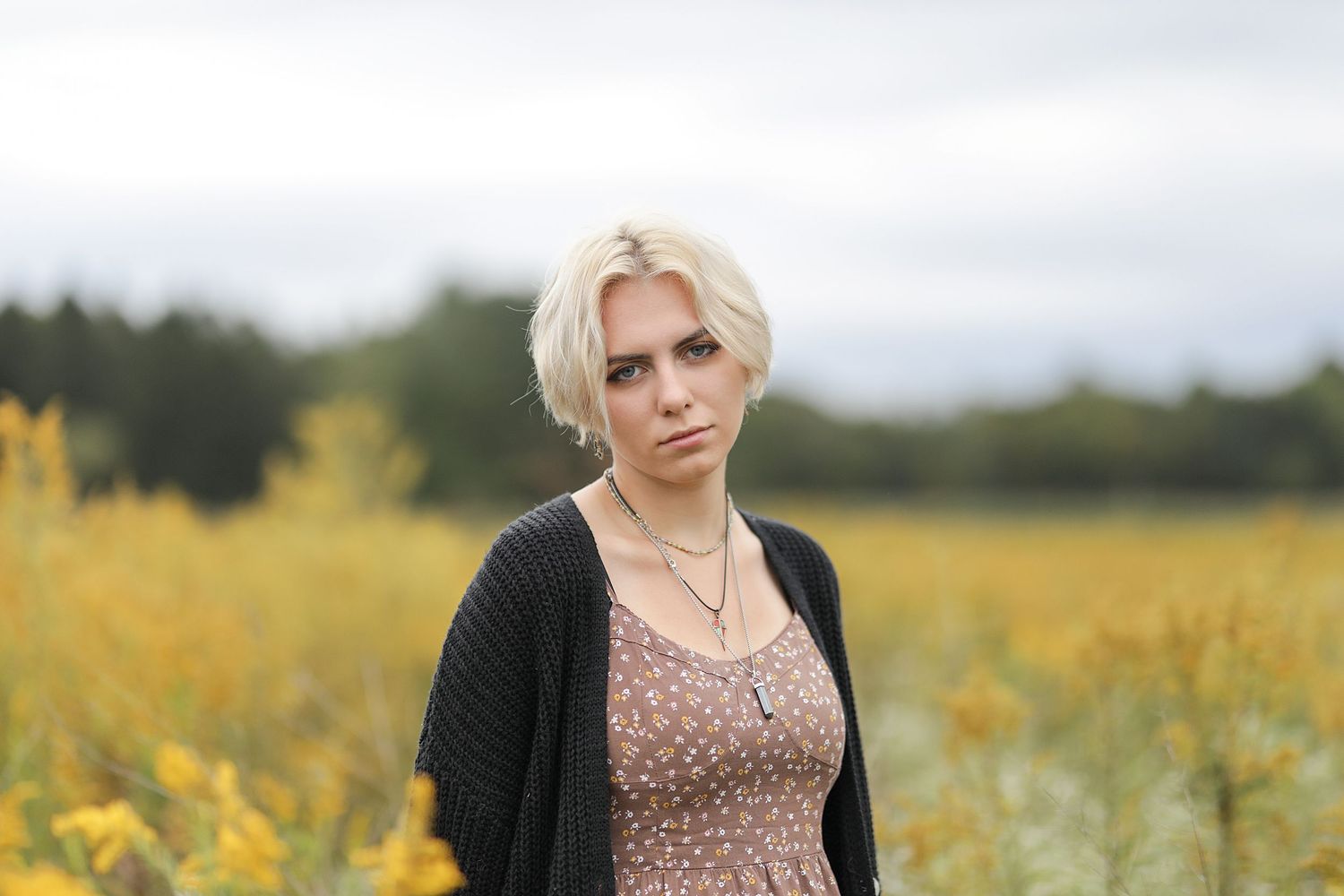 Person in black cardigan and patterned dress stands in field of yellow wildflowers on a cloudy day.