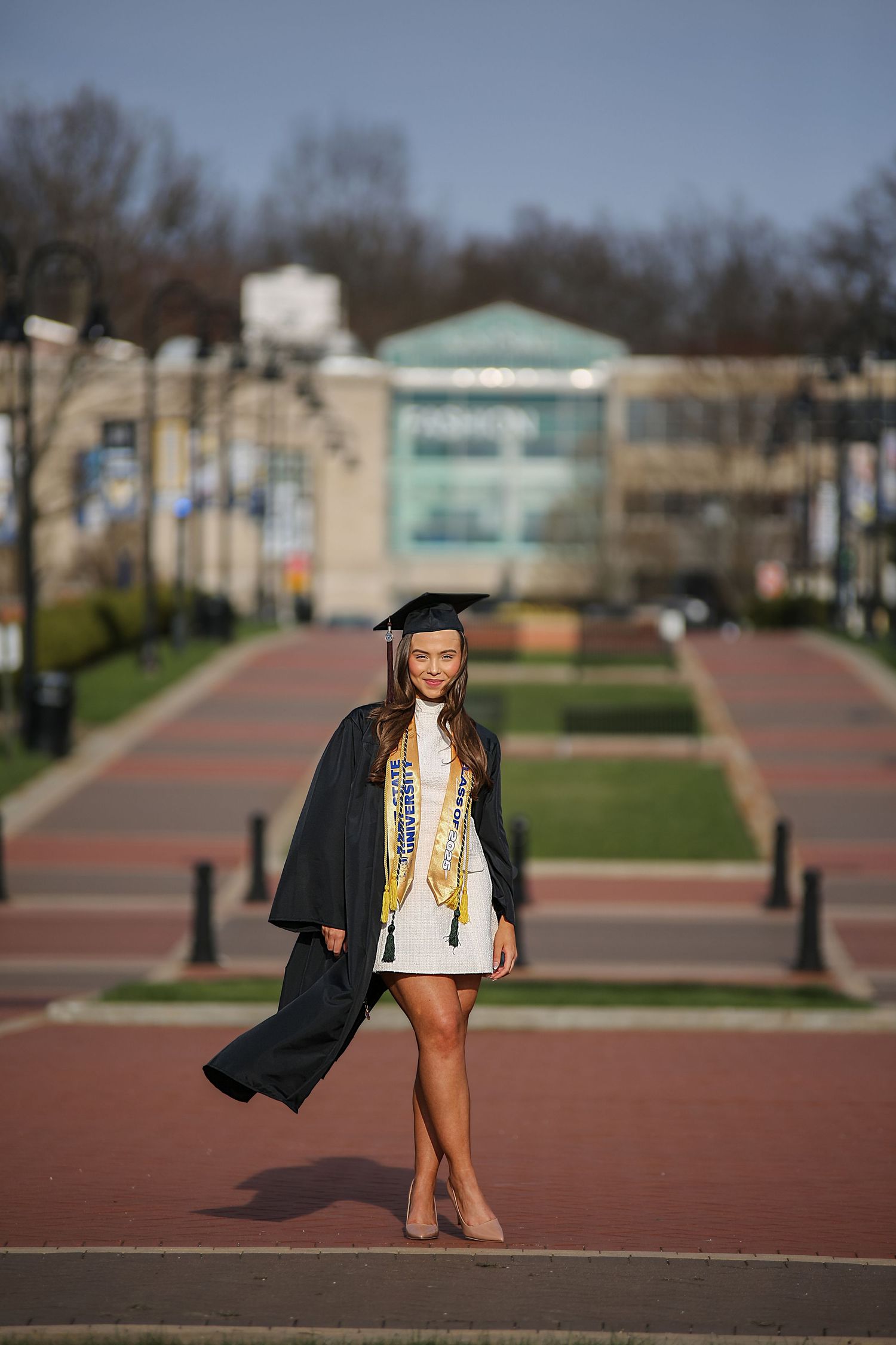 Graduate poses in cap and gown on a brick pathway with campus buildings in the background during a sunny day.
