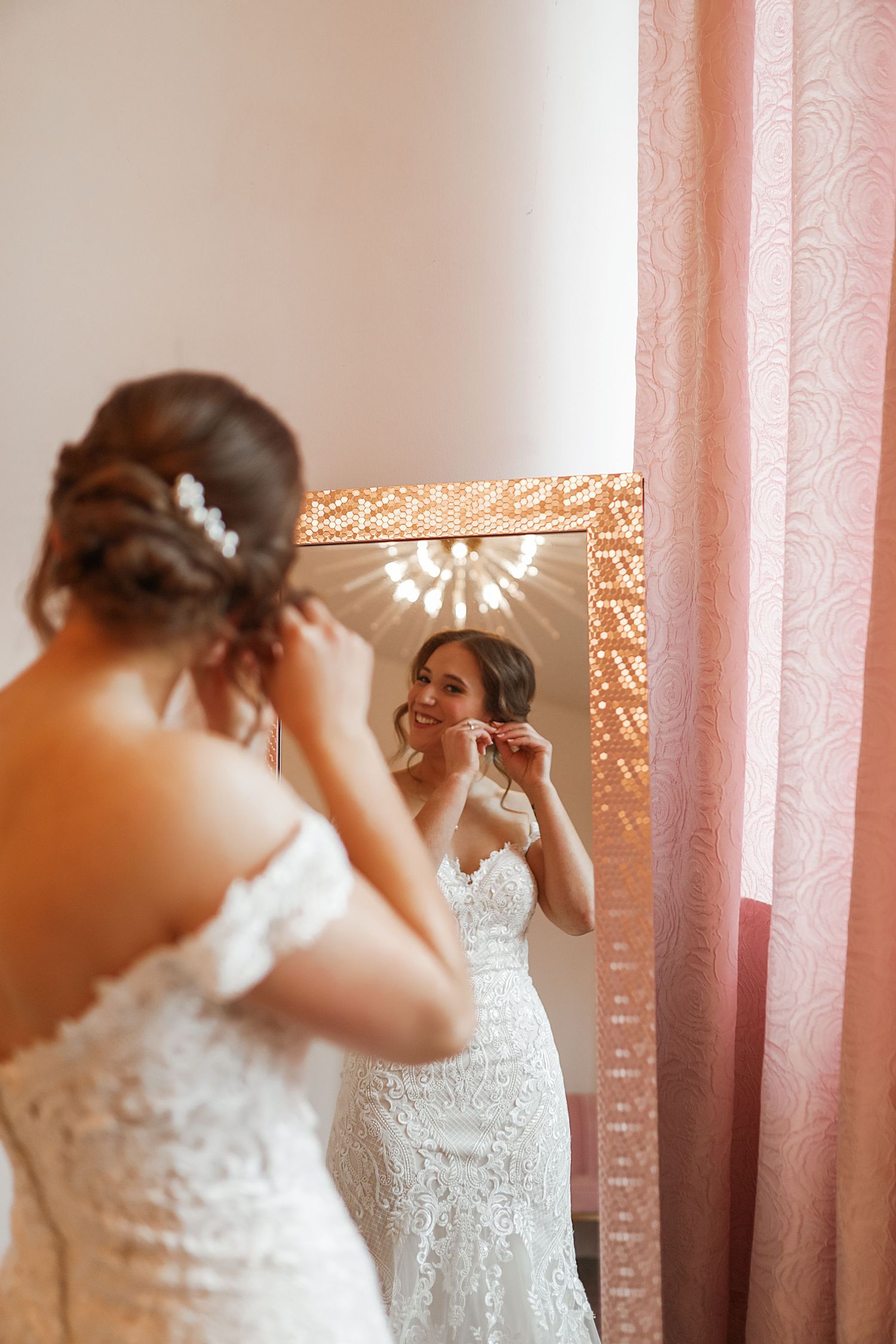A bride in a white lace wedding dress smiles while getting ready in front of a pink curtained window.
