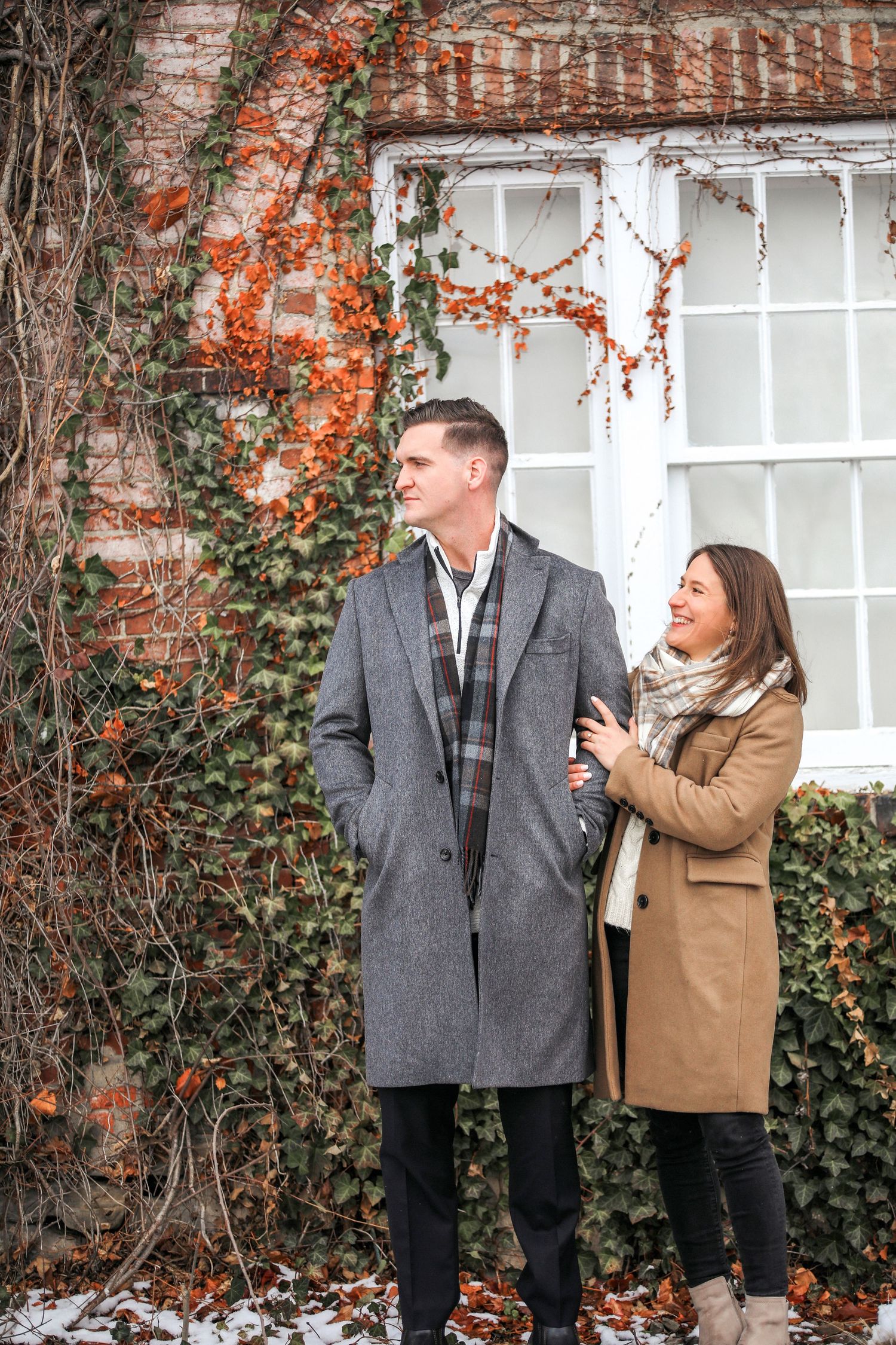 A couple shares a playful moment against an ivy-covered brick wall during a fall photoshoot.