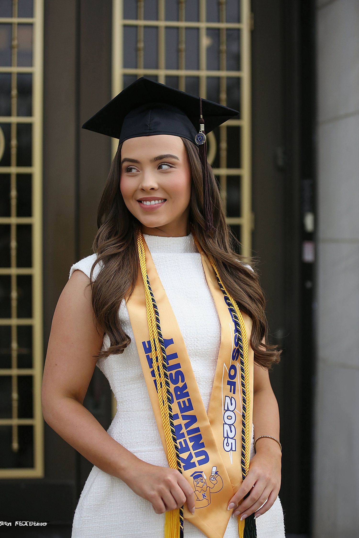 Graduate wearing white dress and gold and blue honor cords poses for graduation photos outdoors.