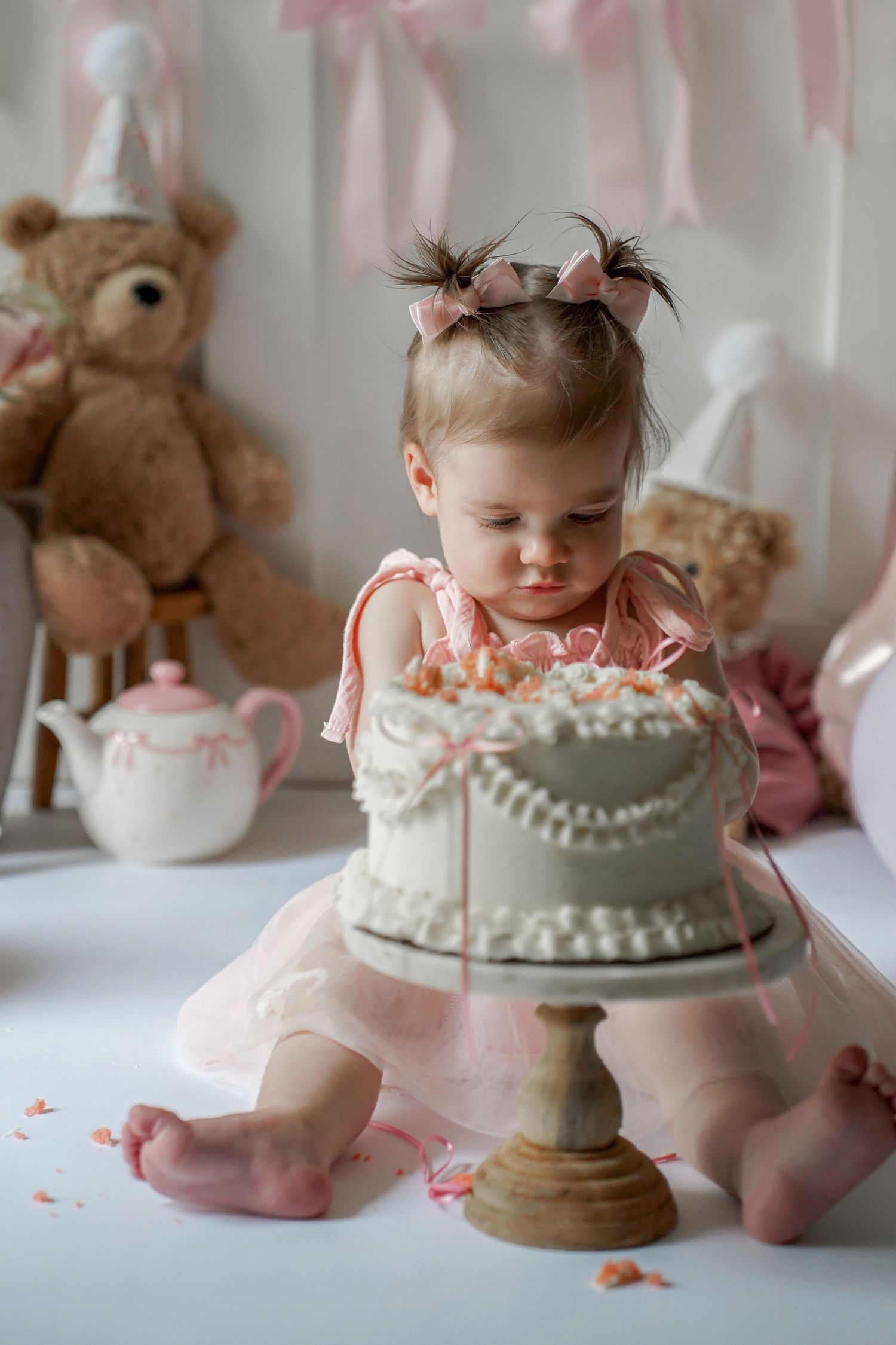 A white birthday cake with pink accents sits on a cake stand surrounded by soft pink decorations and teddy bears.