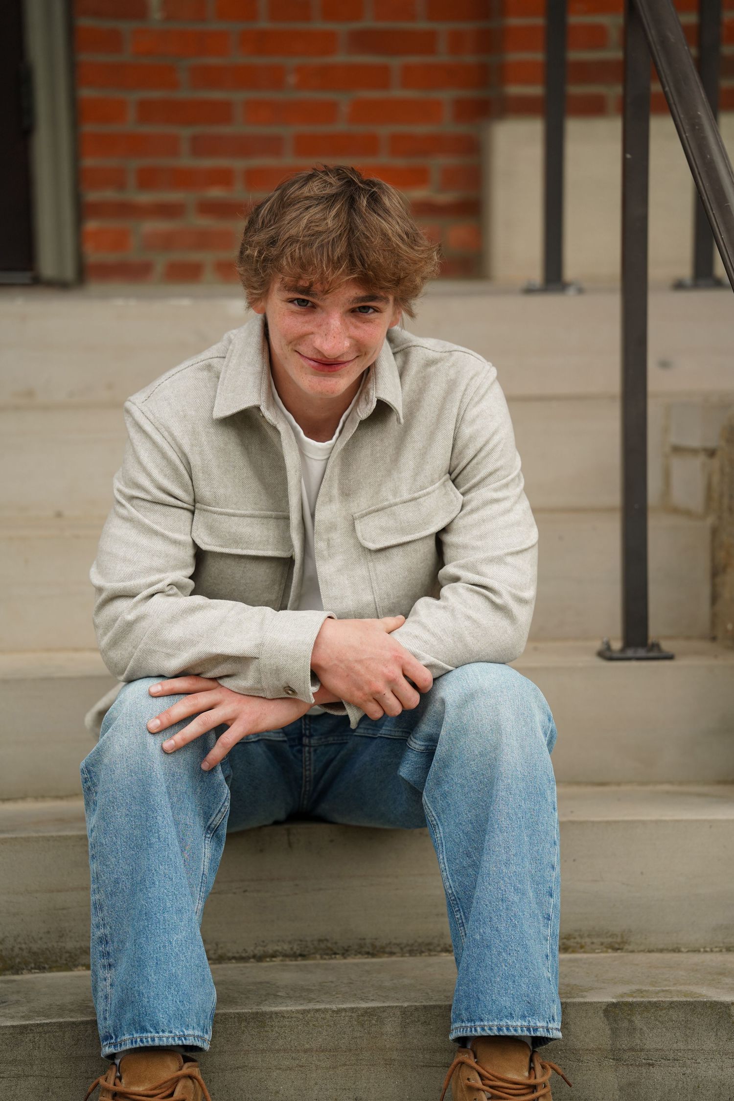 Person in light jacket and blue jeans sits casually on concrete steps against red brick wall.