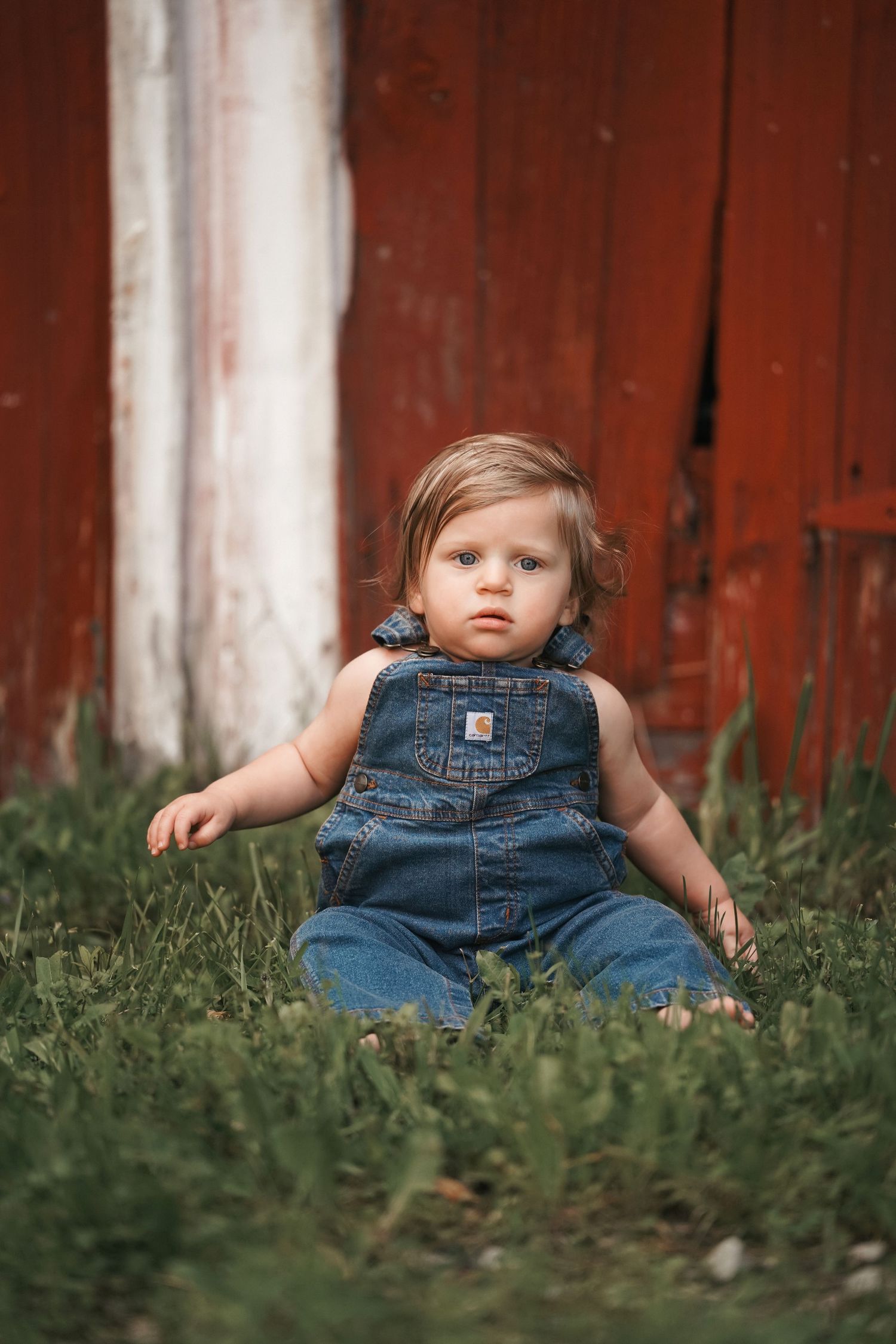 A young child wearing Carhartt denim overalls sits in green grass against a rustic red barn wall.