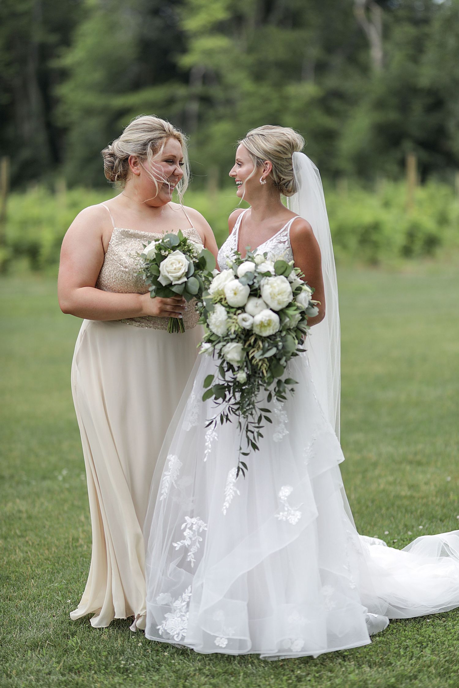 A bride and bridesmaid pose together outdoors holding elegant white floral bouquets.