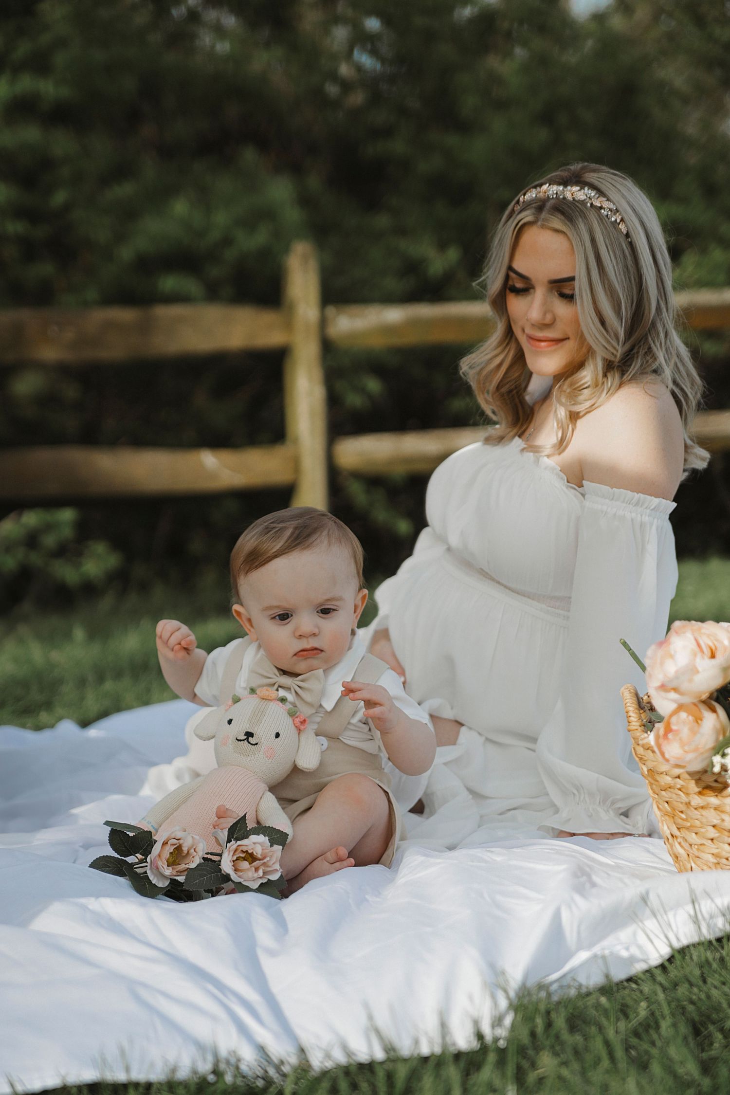 Mother and baby sitting on white blanket having outdoor photoshoot with rustic fence and stuffed toy.
