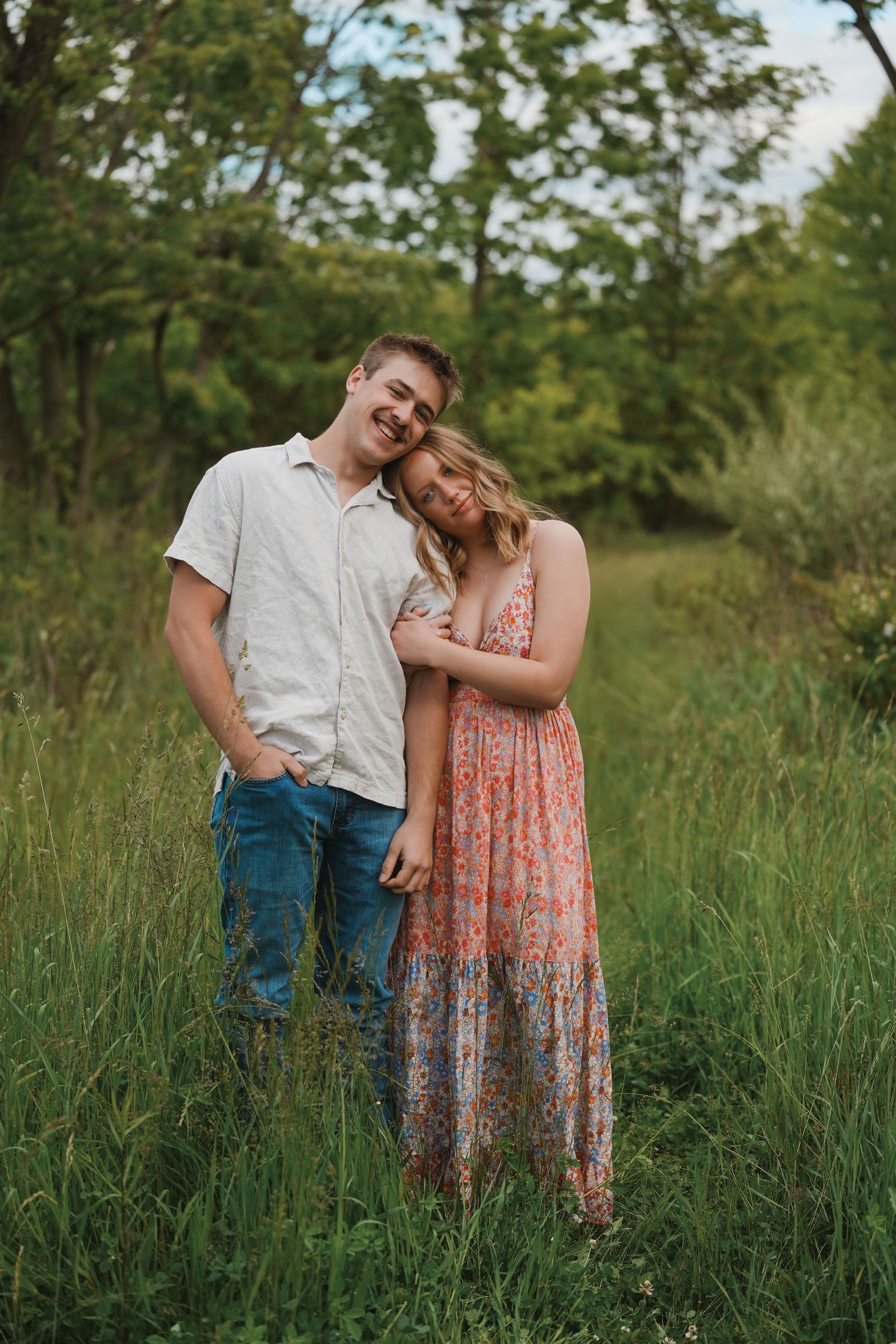 A couple stands together in a grassy meadow on a summer evening, wearing casual clothing amid greenery and trees.