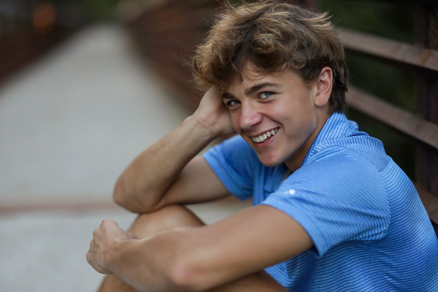 Person in a light blue t-shirt smiling and sitting outdoors with hand near face.