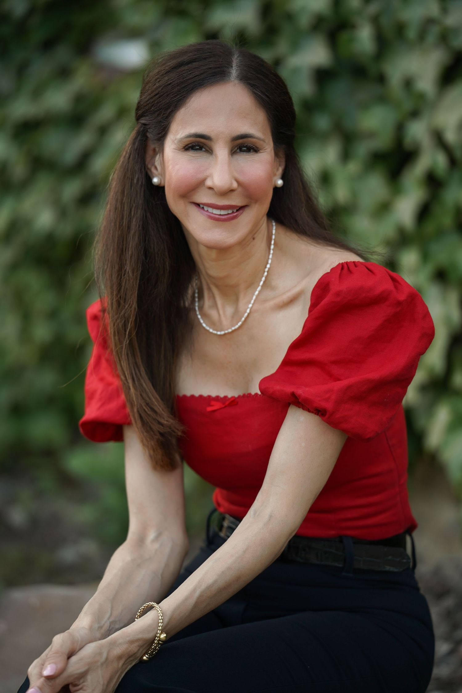 A woman in a red puff sleeve top and black skirt sits outdoors wearing a pearl necklace against a green leafy background.