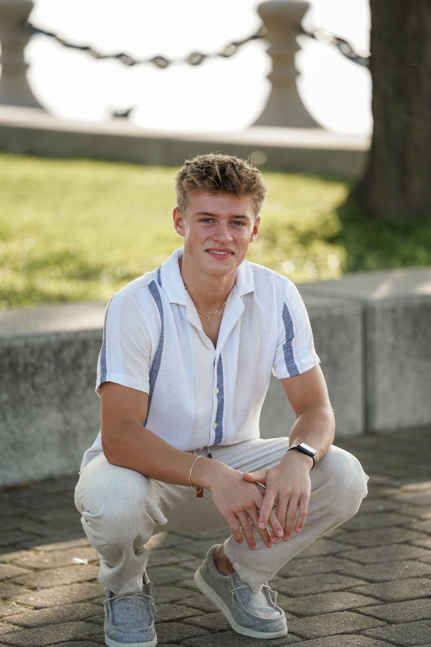 Young person in white shirt and light pants crouching casually on a stone walkway during a sunny afternoon.