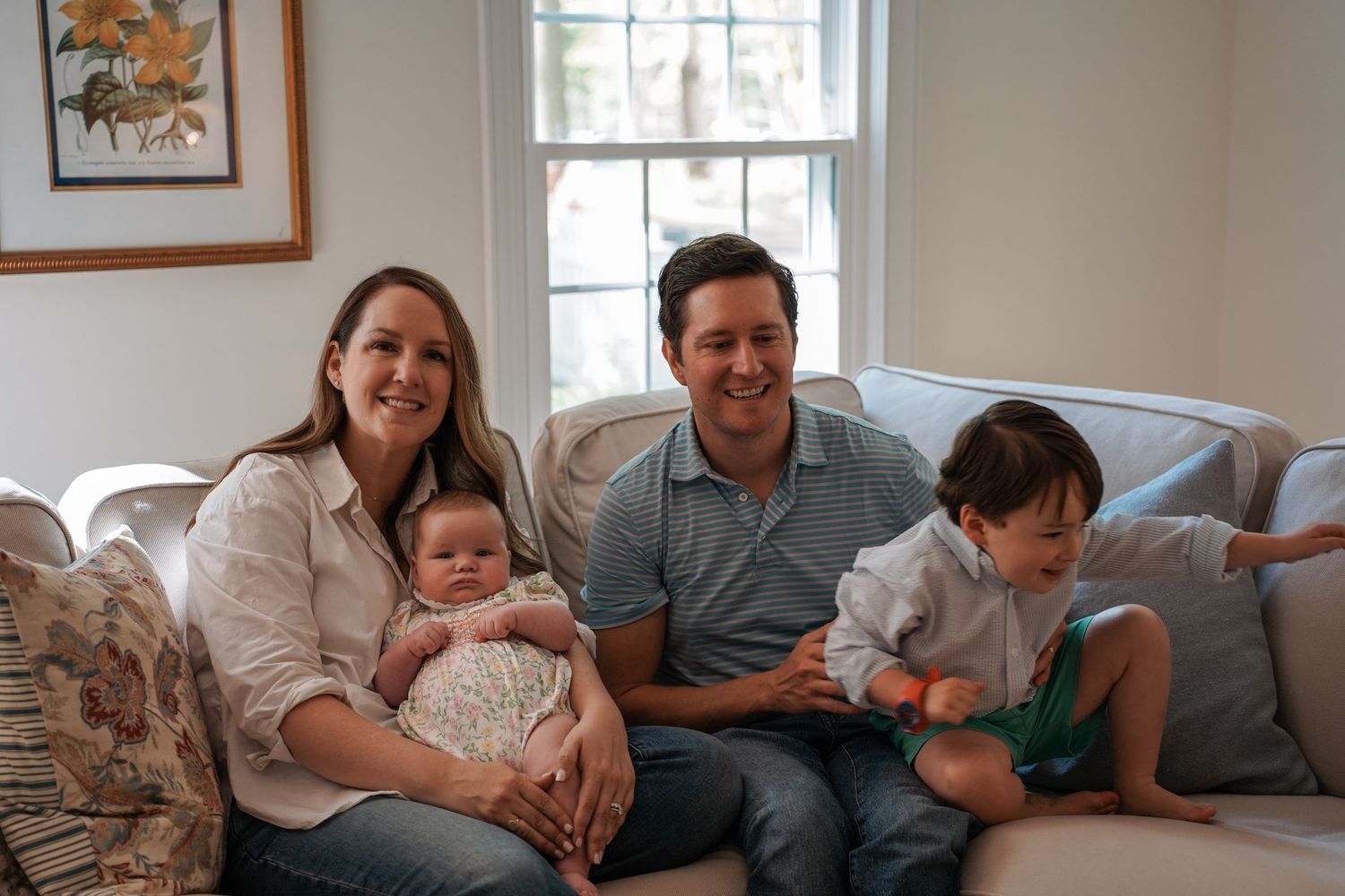 Family relaxing together on a gray couch in a bright living room with a window and framed artwork.