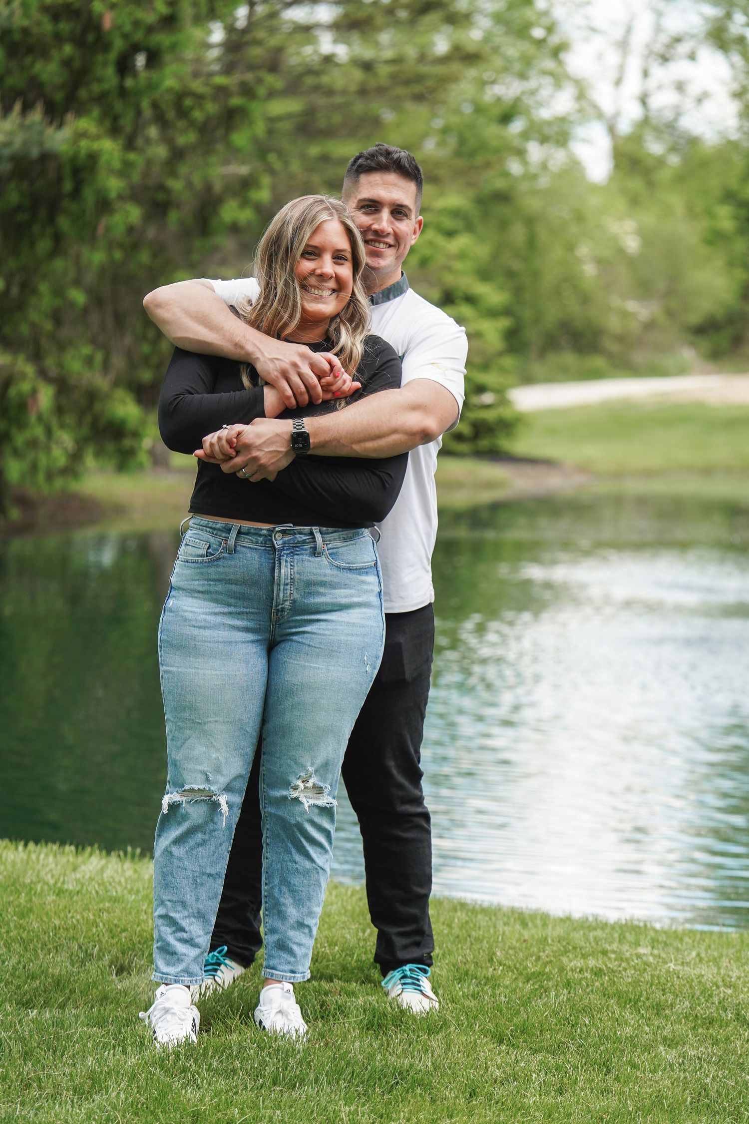 A couple embraces on green grass near a tranquil pond, capturing a sweet romantic moment in nature.