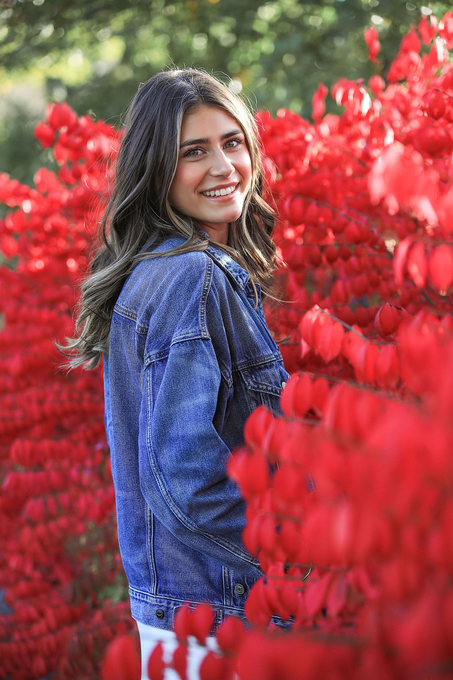 A person in a denim jacket poses outdoors against vibrant red autumn foliage.