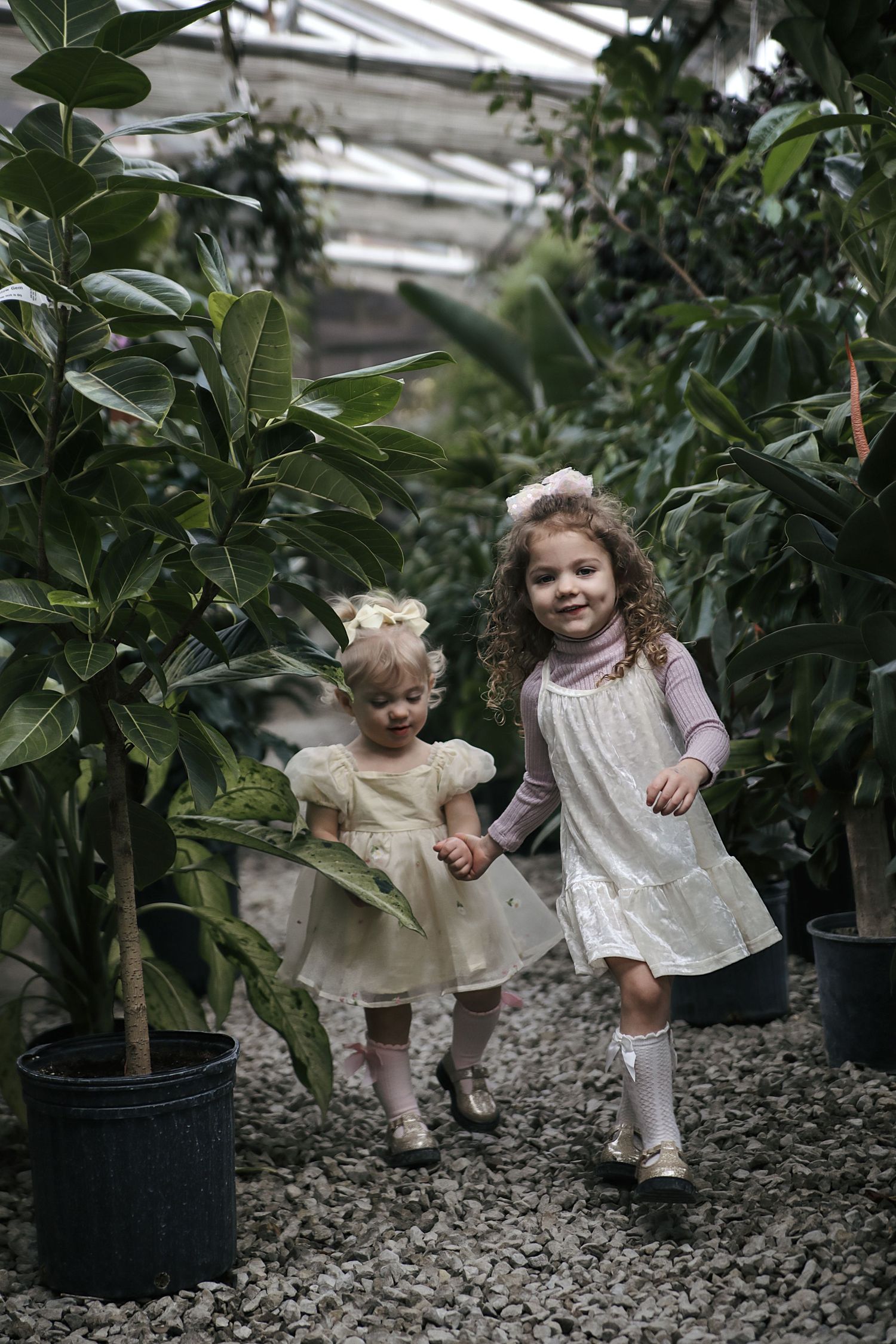 Two children in white dresses walk hand in hand between potted plants in a greenhouse setting.