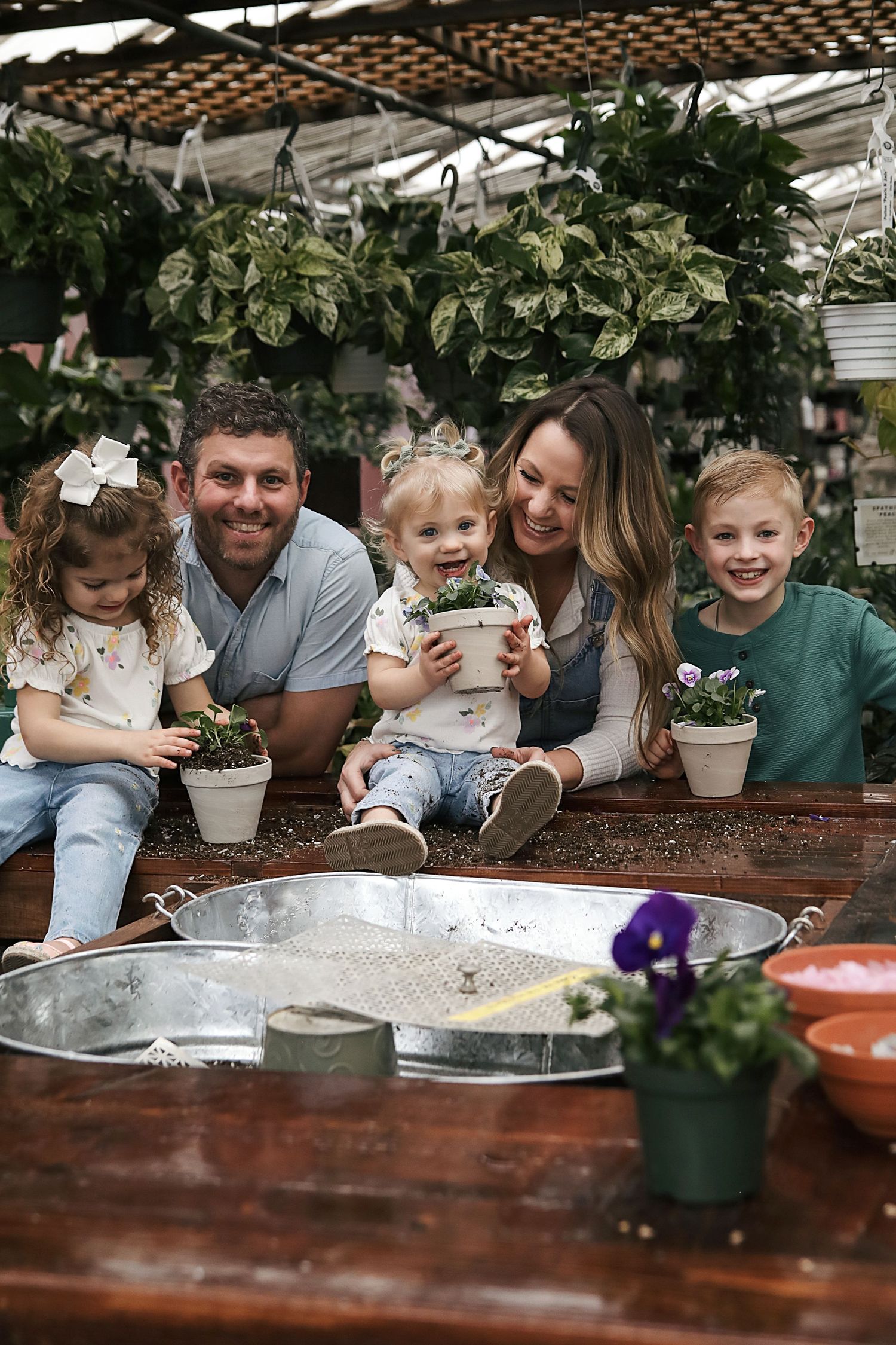 Family enjoying a greenhouse gardening activity together with potted plants and soil on a wooden table.