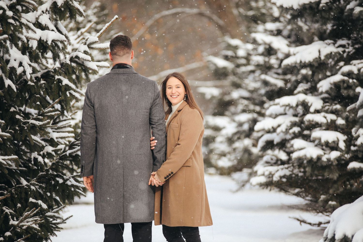 A couple in winter coats walks along a snowy path surrounded by evergreen trees covered in fresh snow.